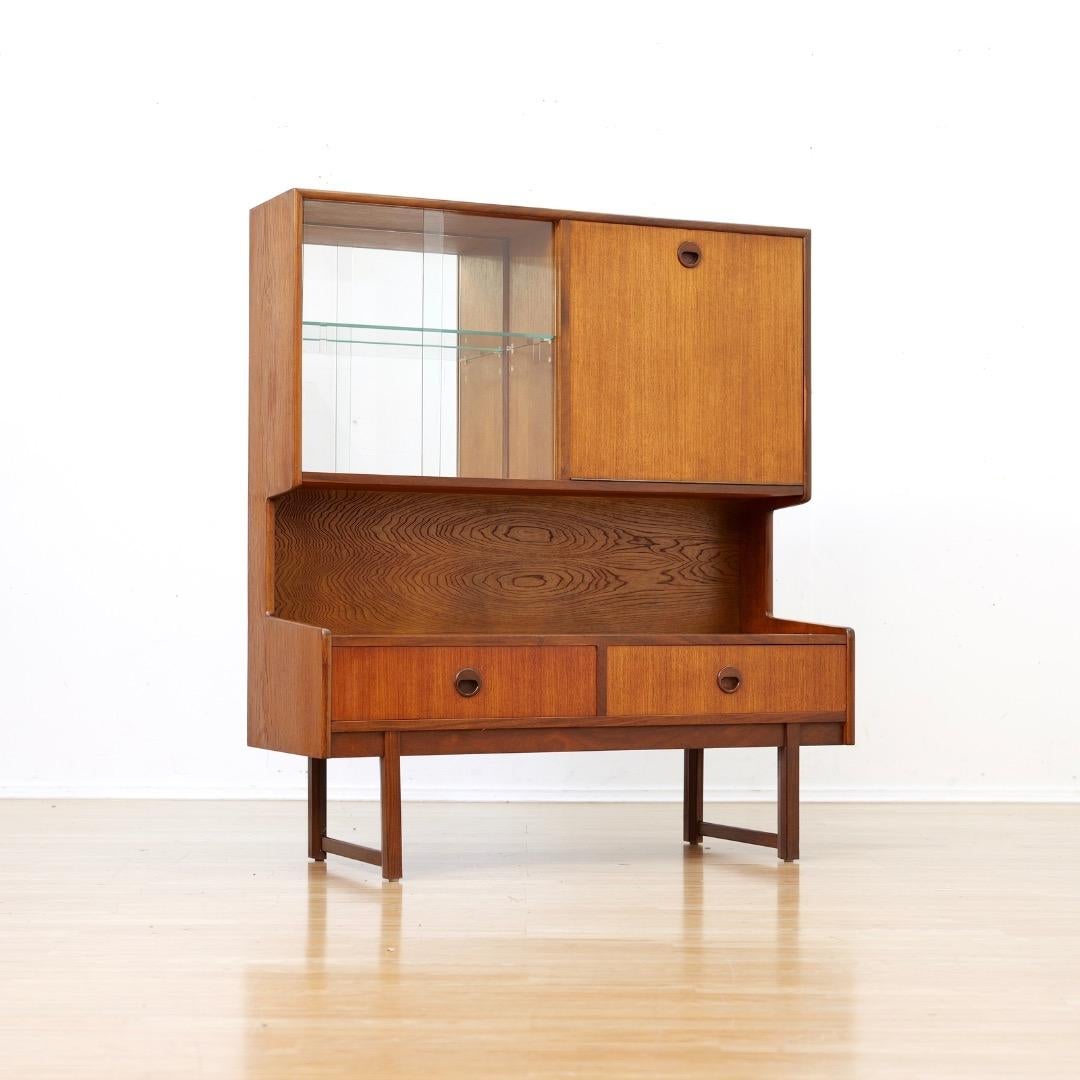 Questa vetrinetta vintage è stata prodotta in Inghilterra da Turnidge of London negli anni Settanta.  Un mobile contenitore dal design meraviglioso, con un portabottiglie/bicchieri nella parte superiore, un ripiano centrale e cassetti inferiori, è