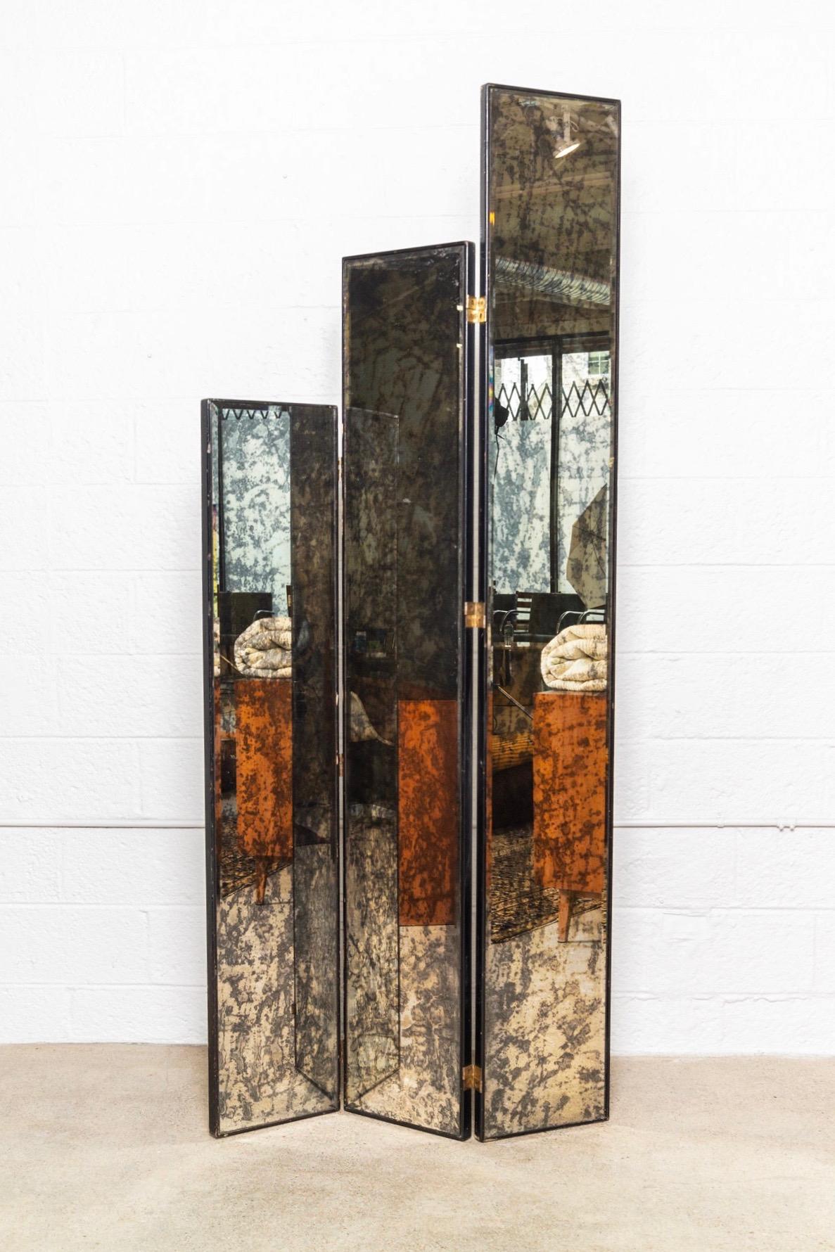 This stunning vintage midcentury folding mirrored room divider is circa 1970. It features three tall double sided panels of antiqued smoked mirror with an 1/2” beveled edge in a thick black wooden frame. Mirrored panels are on brass hinges and can