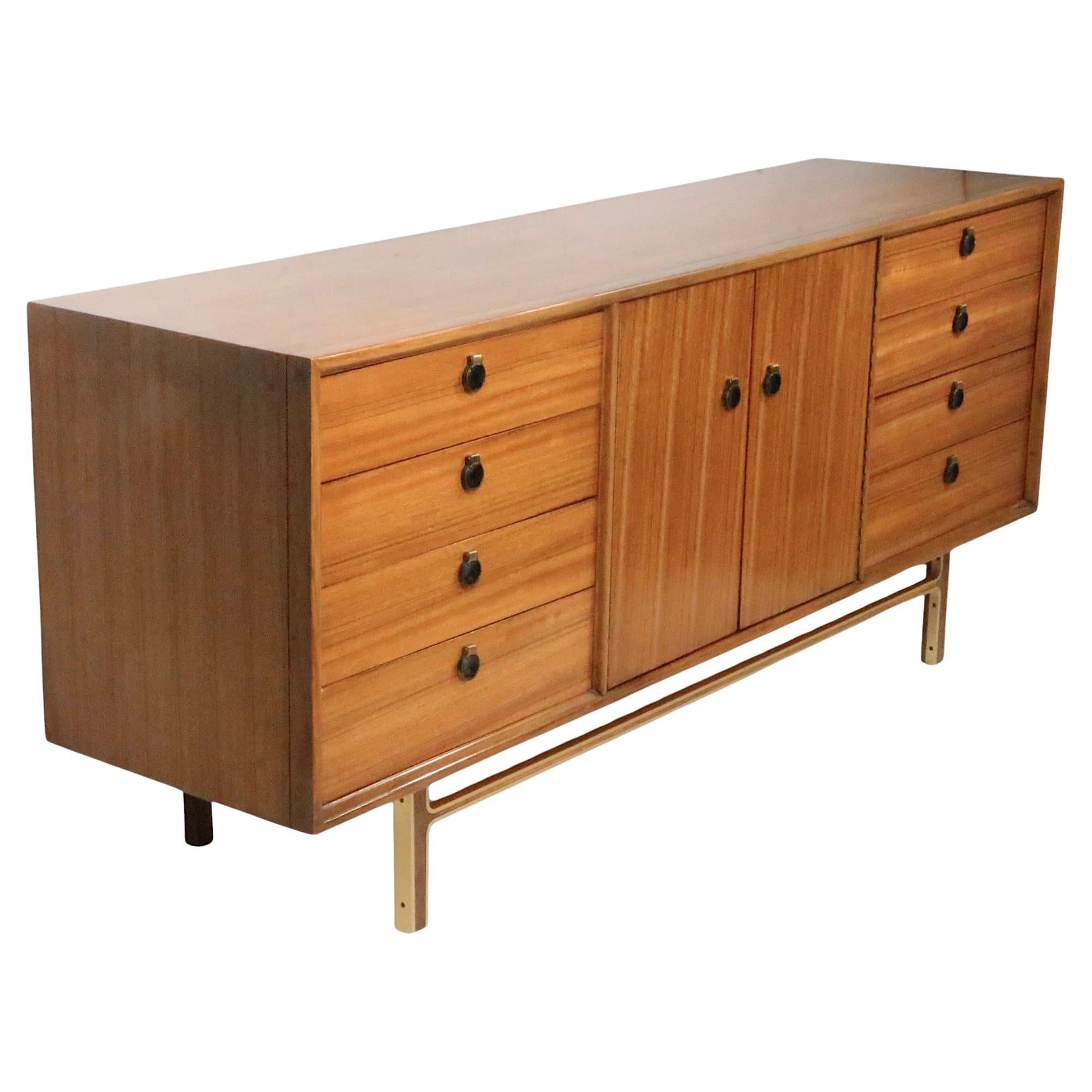 Chic long low mid century dresser with two banks of four drawers, flanking two doors which open to reveal an addition four interior drawers. The rectangular wood case is supported by stylized wood legs, with gold toned anodized aluminum trim. 
This