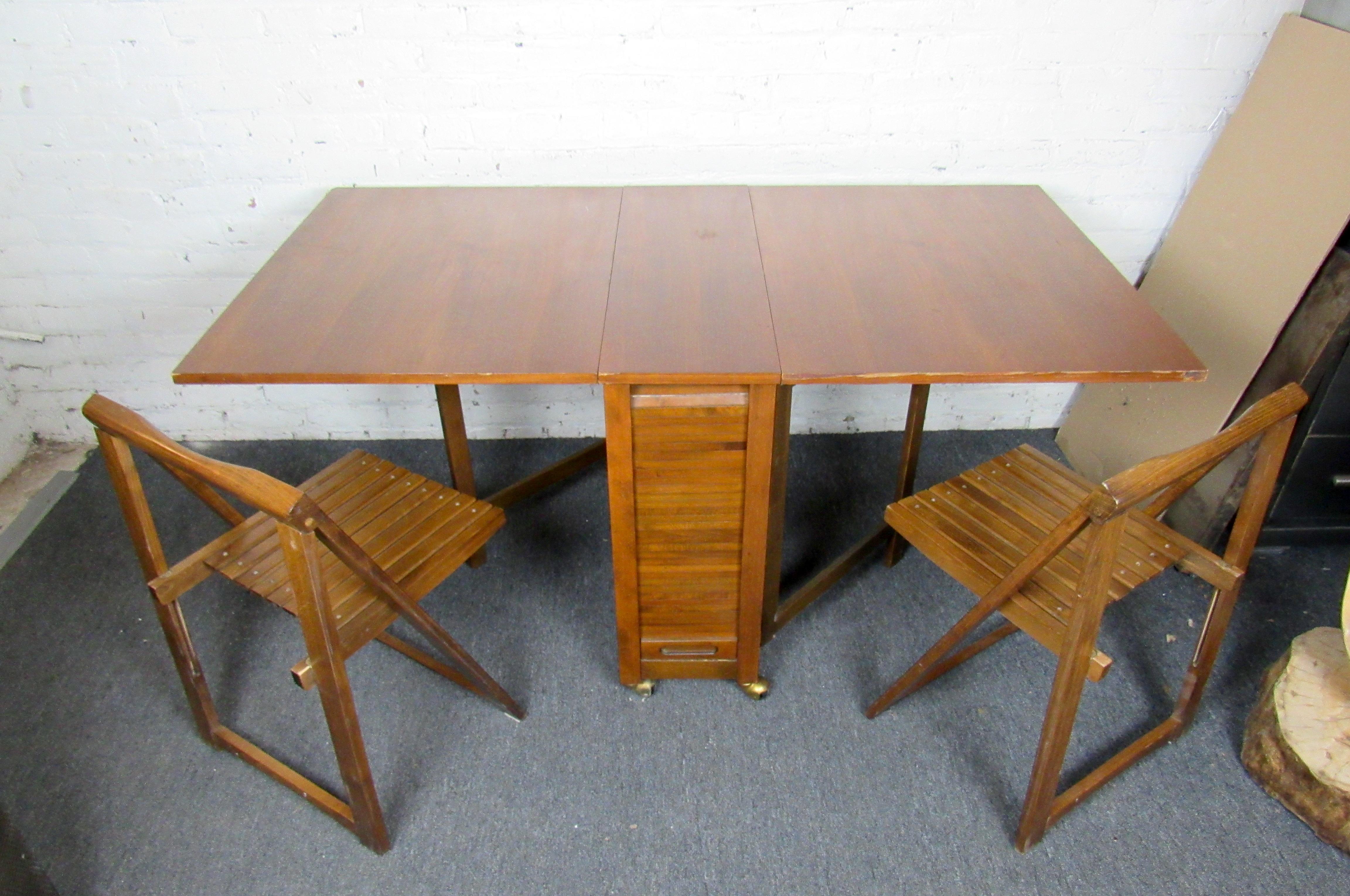 A timeless Mid-Century Modern design, this drop leaf table stores away in a compact design with storage for four folding chairs. Rich woodgrain is complemented by brass accents on the table's rolling base. Please confirm item location with seller