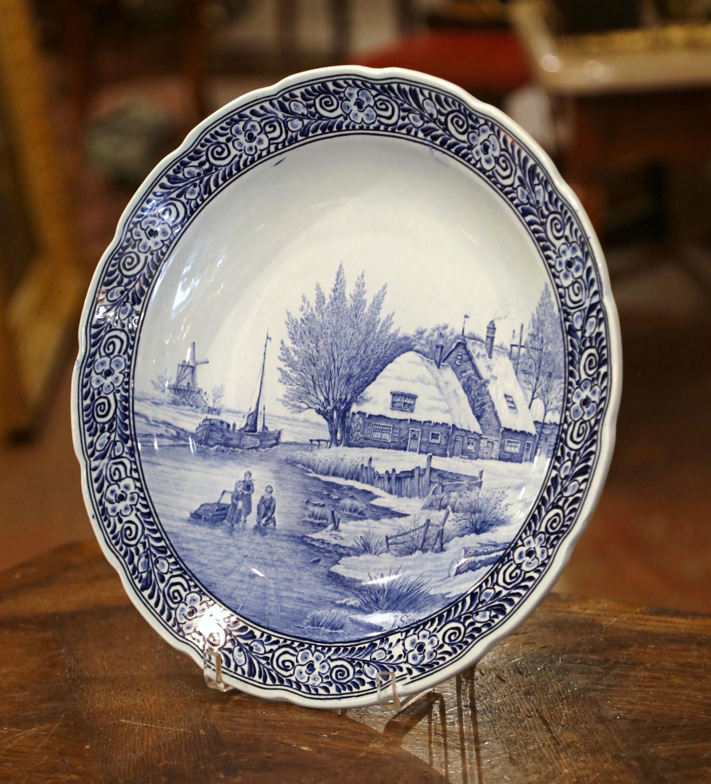 This large antique charger was crafted in Holland, circa 1960. The large, wall hanging plate depicts a wintry cottage scene, a Dutch windmill and boat, as well as people enjoying the frozen lake! One person is assembling skate shoes, while the other