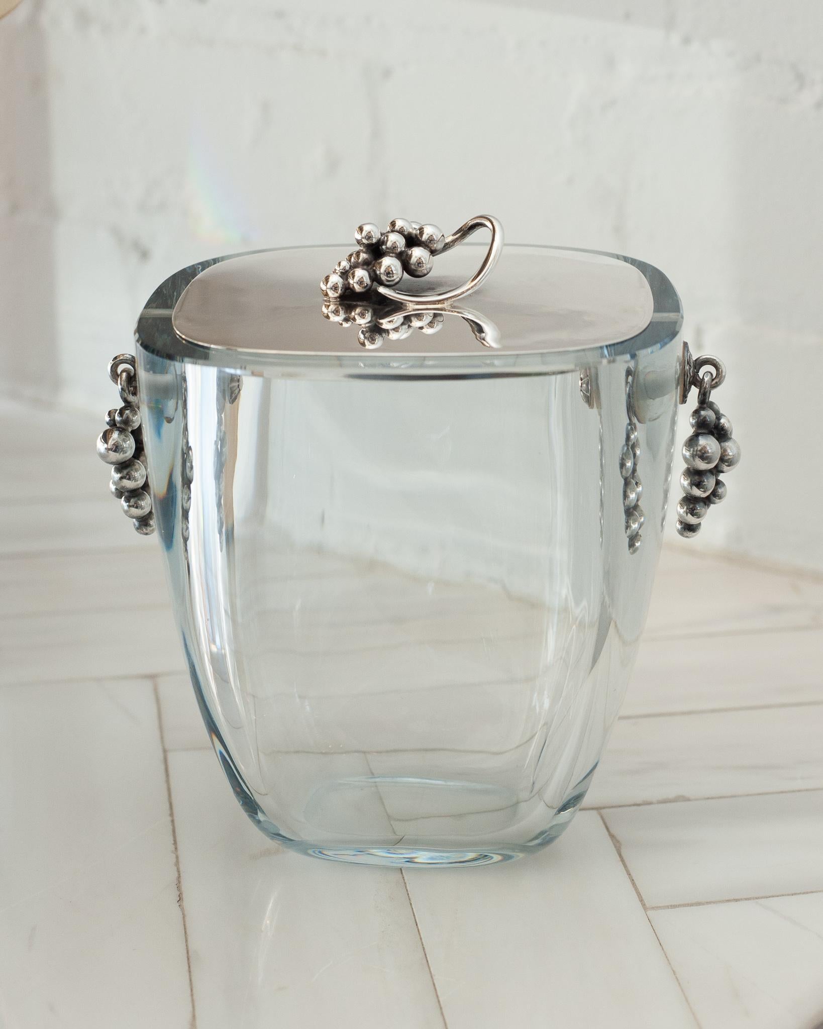 A beautiful mid century ice bucket made by E. Dragsted in Denmark. It features a thickly cut oval clear bucket, with sterling silver grape bunches mounted to each side. Flat sterling silver cover with grape bunch finial. Cover marked with makers