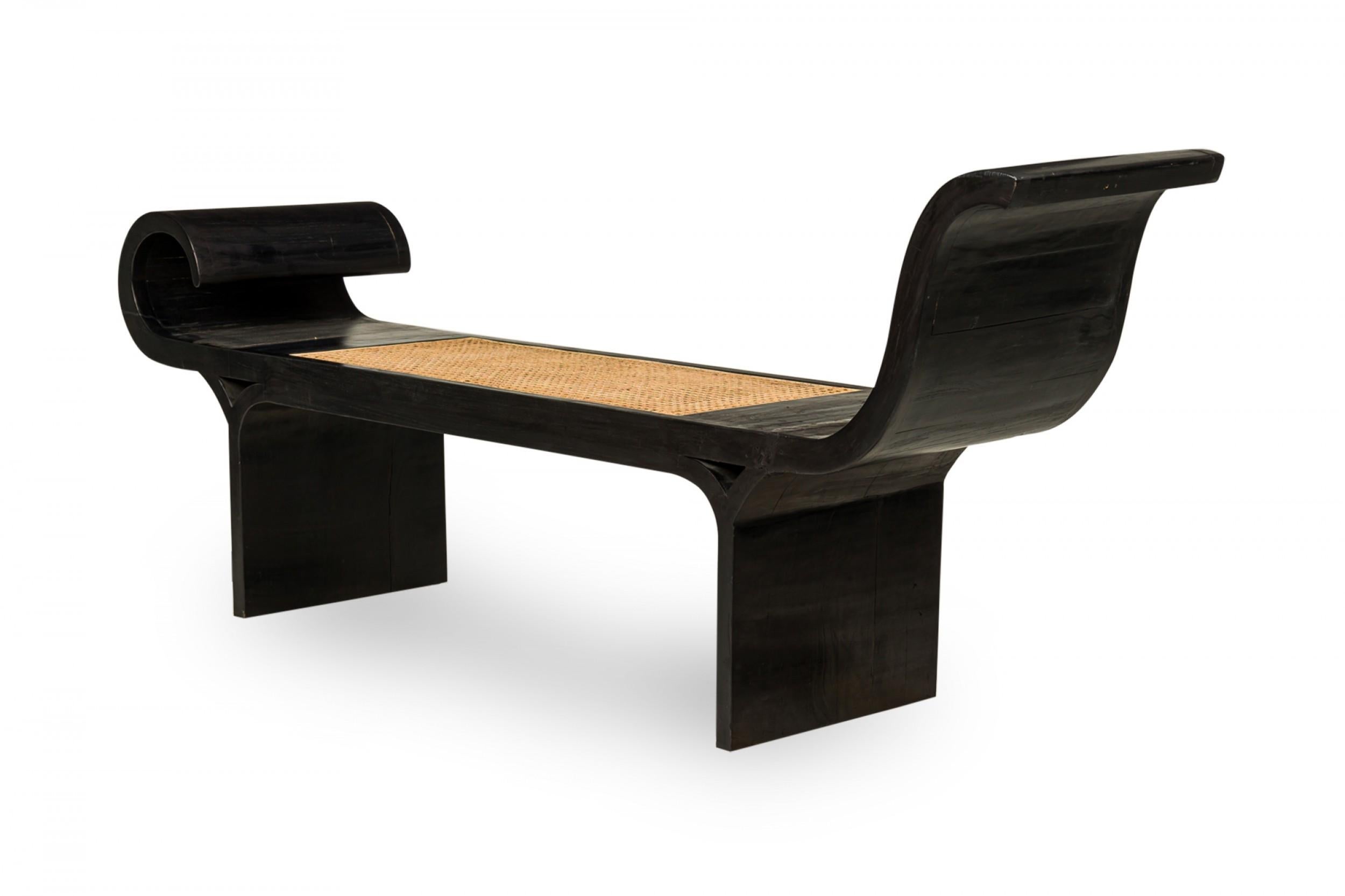 Banco de madera ebonizada de mediados de siglo, con dos laterales de volutas y un asiento central de caña, que descansa sobre dos patas rectangulares.

Estado: Bueno; Desgaste acorde con la edad y el uso