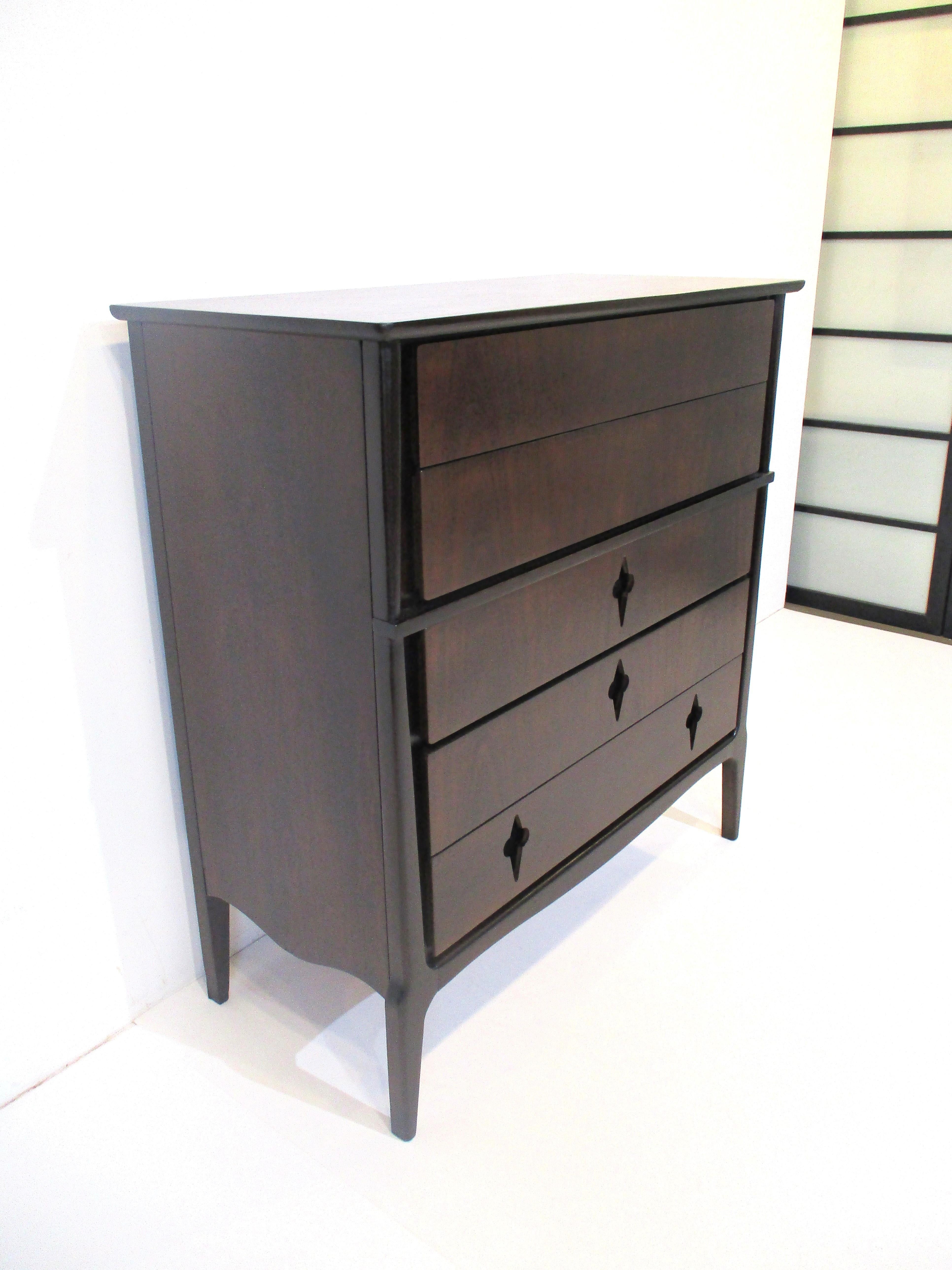 A ebony toned walnut five drawer dresser chest with darker contrasting wood frame giving the piece a rich and well crafted look . The three bottom drawers have a de-lis fleur midcentury styled cutouts used for pulls, manufactured by the United