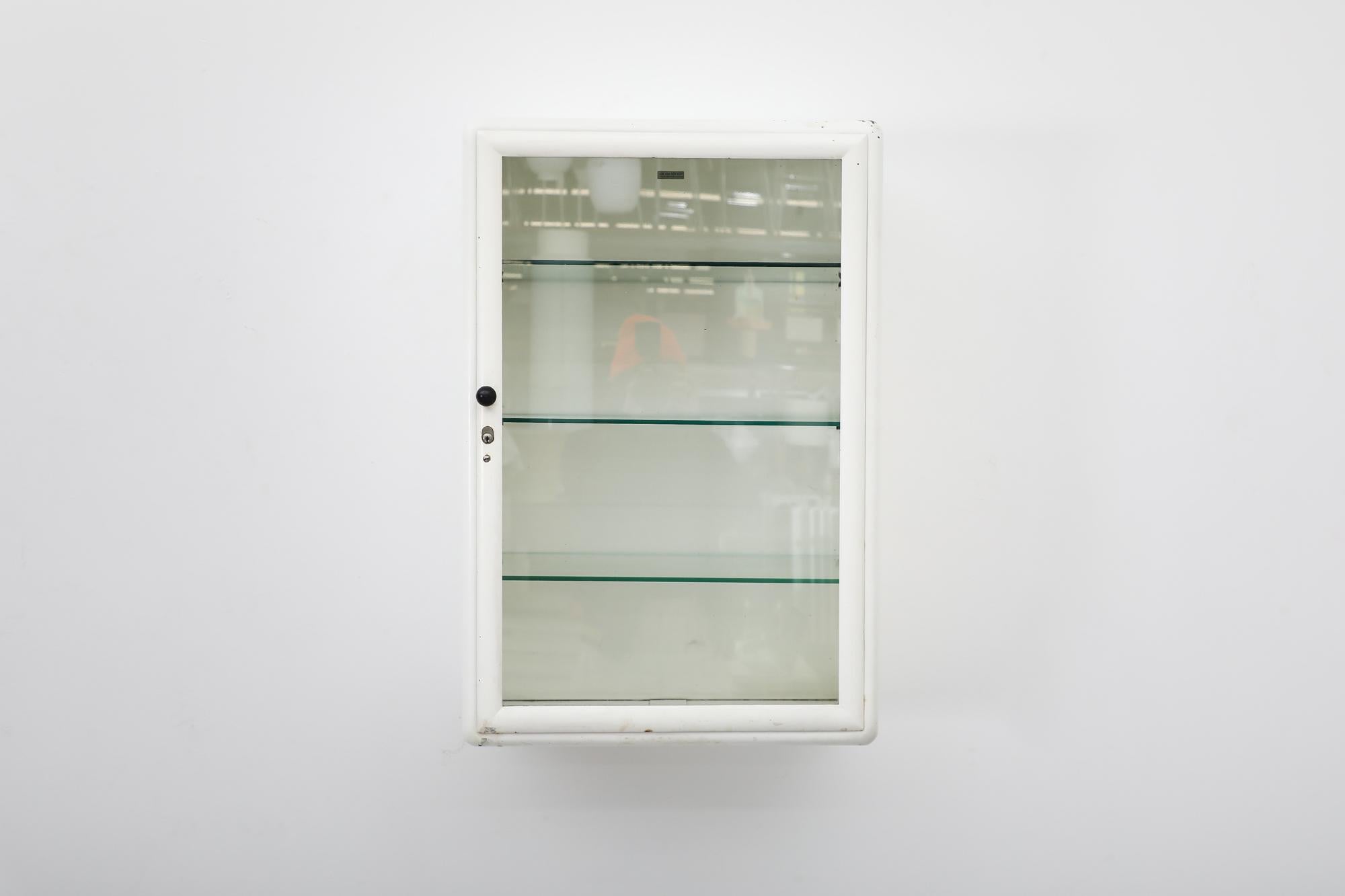 Vintage white enameled metal wall mounted medicine cabinet with glass door and glass shelves. Missing key. In very original condition with visible wear and patina, including scratches and some paint loss. Wear is consistent with its age and use.
