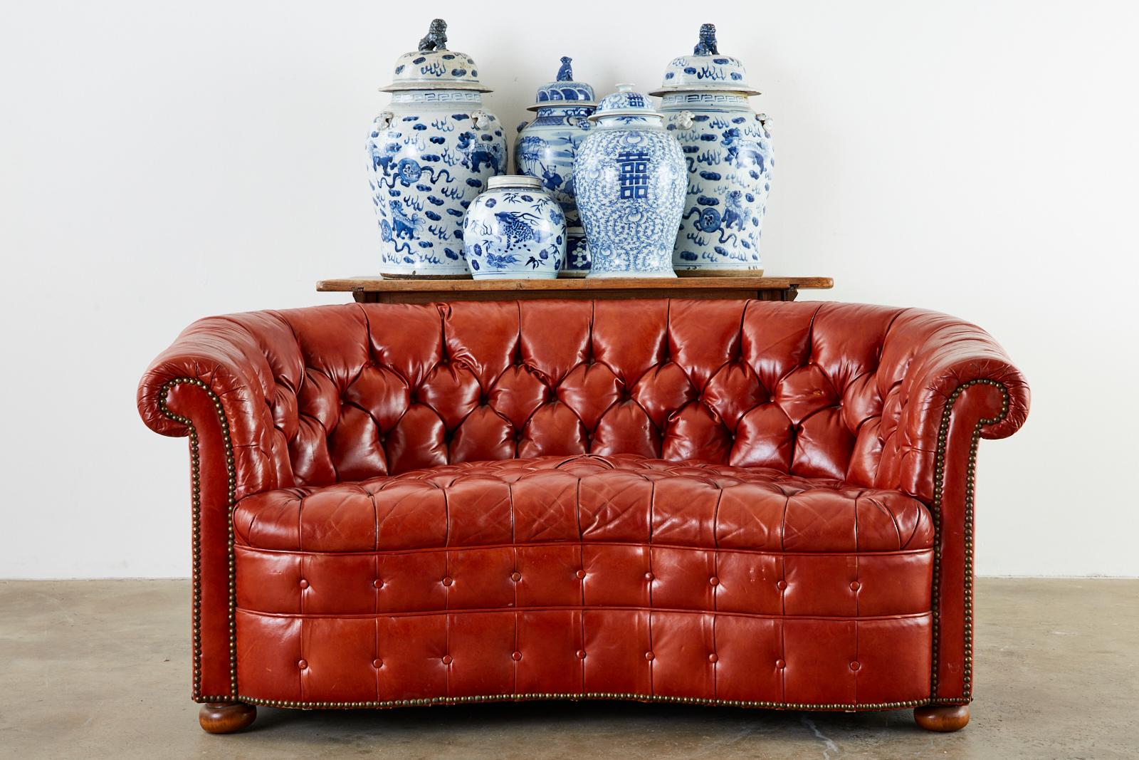 Stylish Mid-Century Modern English Chesterfield settee or loveseat featuring a curved, round back and a serpentine front seat. The kidney style shape has a hardwood frame covered with orange tufted leather hides bordered with brass tack nailheads.