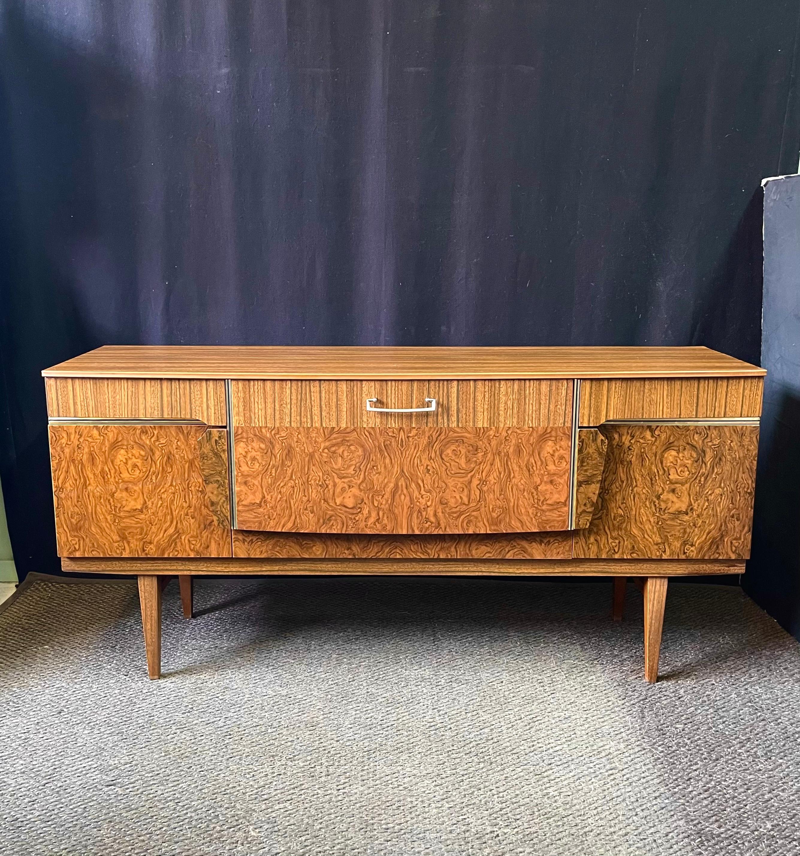 This vintage mid-century credenza was designed and manufactured by 'beautility furniture' in England during the 1950s. A posh, stylishly designed credenza, the burl wood melamine veneered cabinet rests on tapered legs. The case consists of two outer