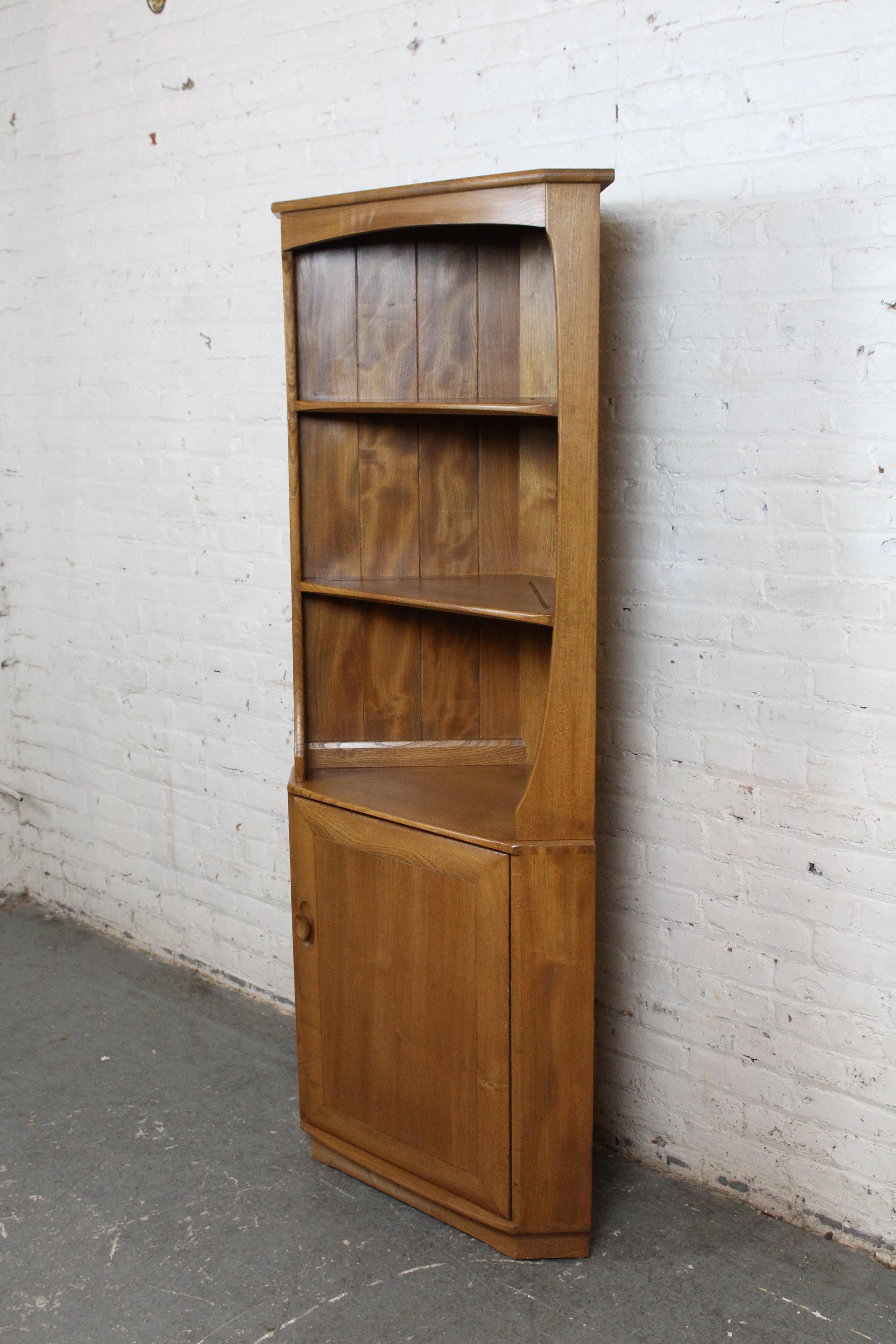 Charmingly chic solid elm triangular corner cabinet manufactured by Ercol of England in the mid-century post-war era. With an impressive 6 foot height, it houses three open shelves atop a single door cabinet concealing additional shelved storage. A