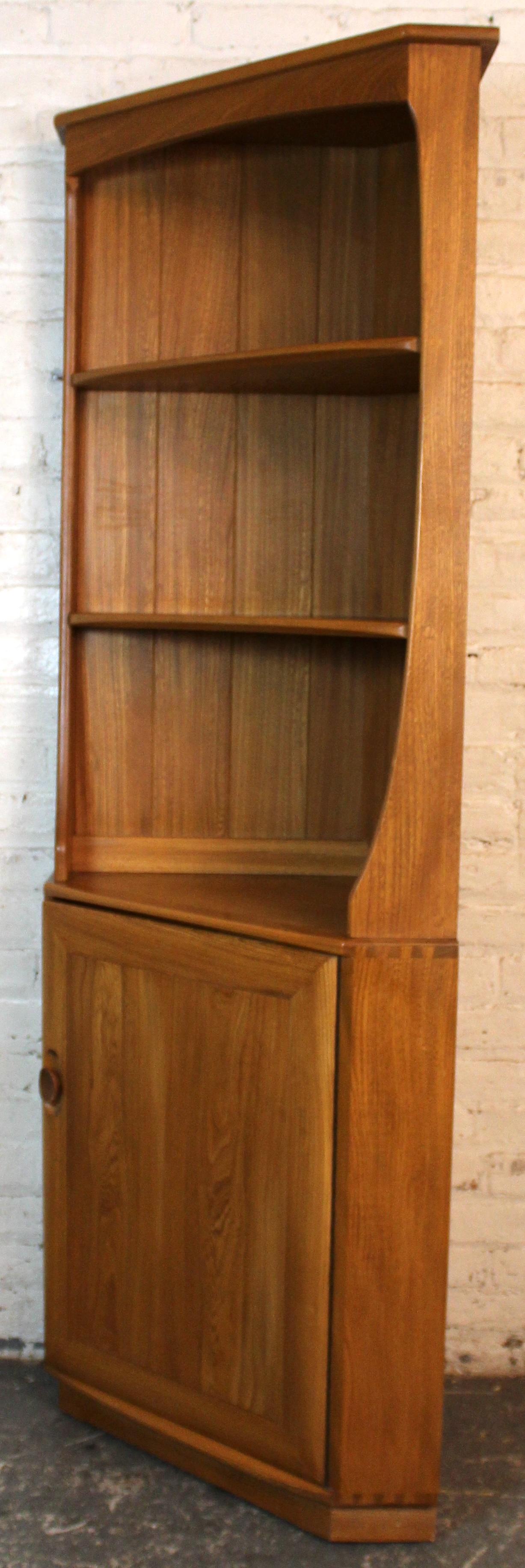 Mid-Century Modern solid elm corner cupboard by English maker Ercol, circa 1950s. With its clean triangular “corner” footprint, the piece features three open display shelves above a single cabinet door concealing additional shelving. Crafted in