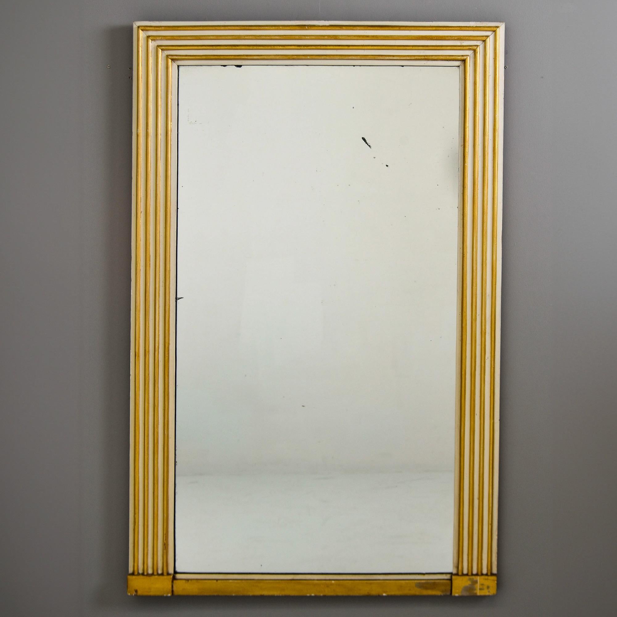 Trouvé en Angleterre, ce miroir à cadre en bois date des années 1950. Le cadre rectangulaire en bois est fini en doré et blanc crème avec un détail de quadruple cannelure en haut et sur les côtés et une finition dorée solide à la base. Le miroir