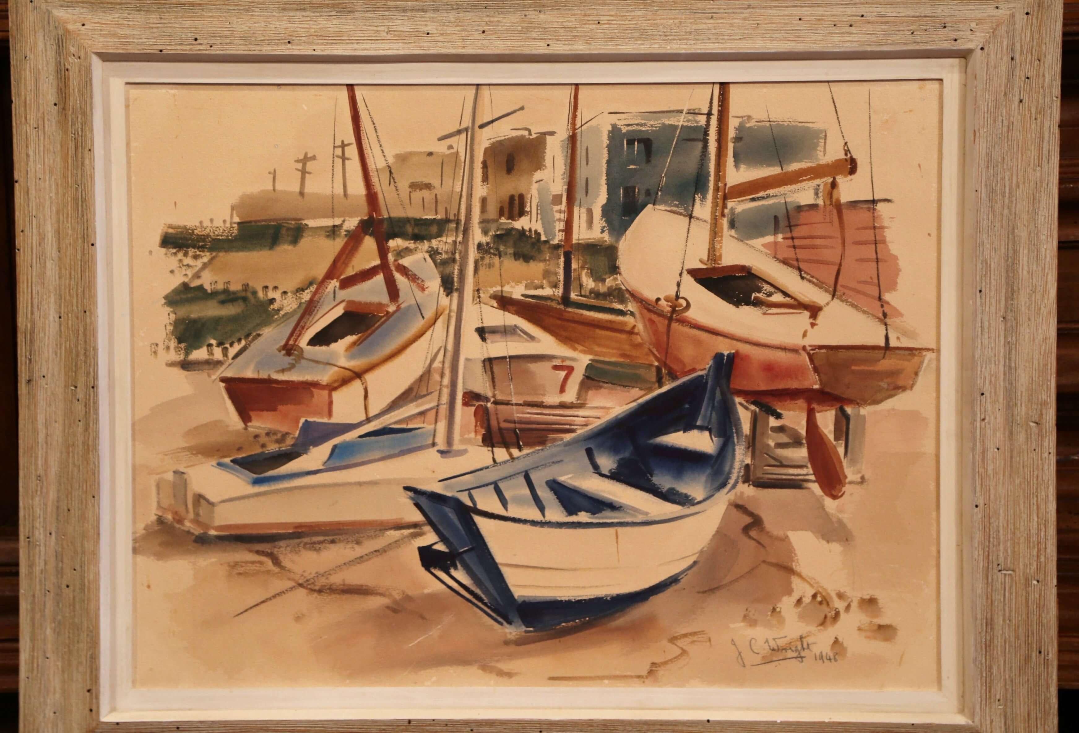 This mid-20th century painting was found in southern France; set in the original wooden frame with white wash finish, the colorful painting features sailboats in harbour with housing in the background. The work of art is signed by the artist J.C.