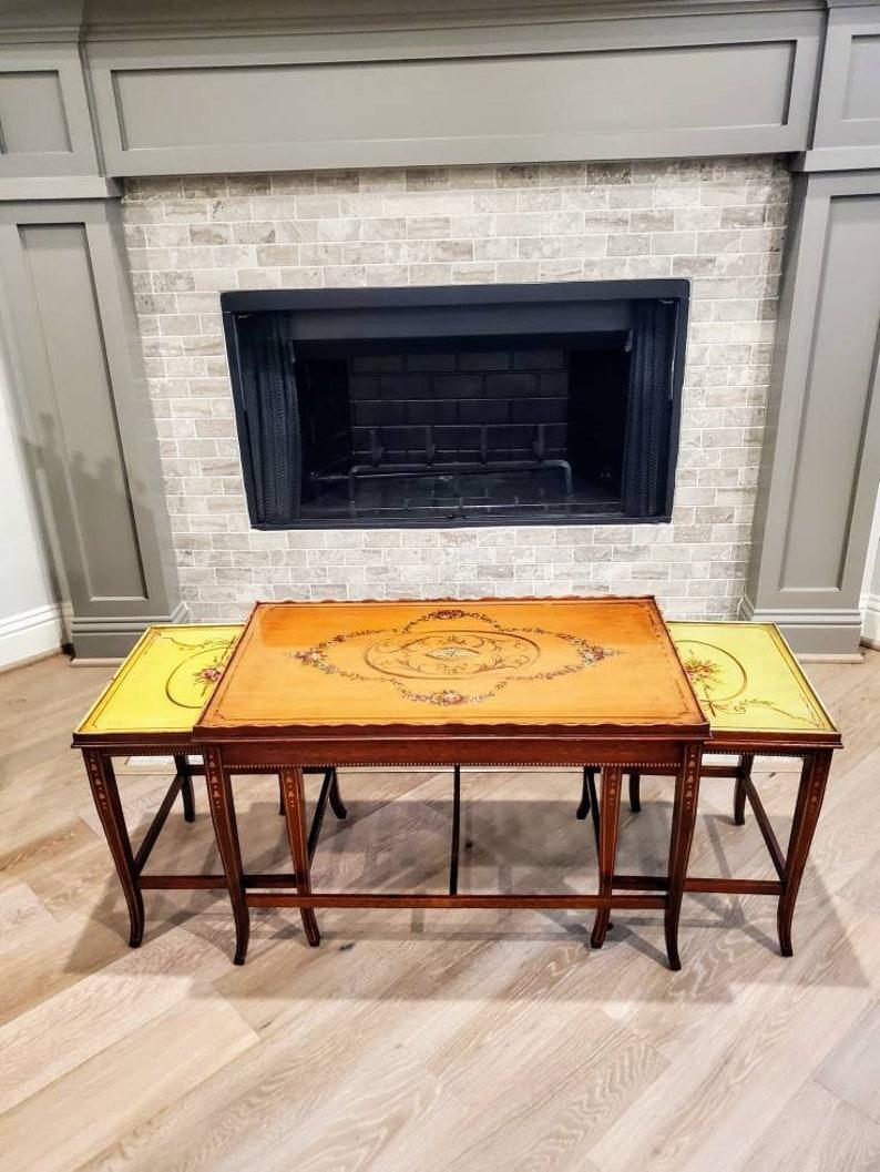 An elegant nest of three English Regency Adam style hand painted mahogany tables. Born in the mid 20th century, the main table (coffee or cocktail table), having rectangular top with undulating shaped raised gallery, framing a removable inset glass