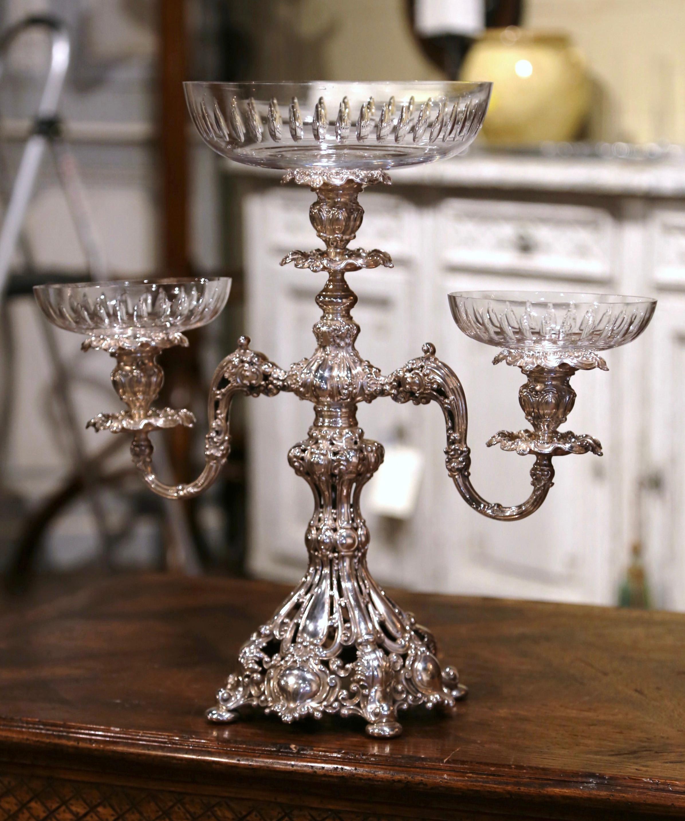 This elegant copper silver plated epergne was created in England, circa 1960. The antique centerpiece stands on rounded feet over a bottom dome decorated with intricate motifs. The dish with two arms features a large center cut crystal bowl and two