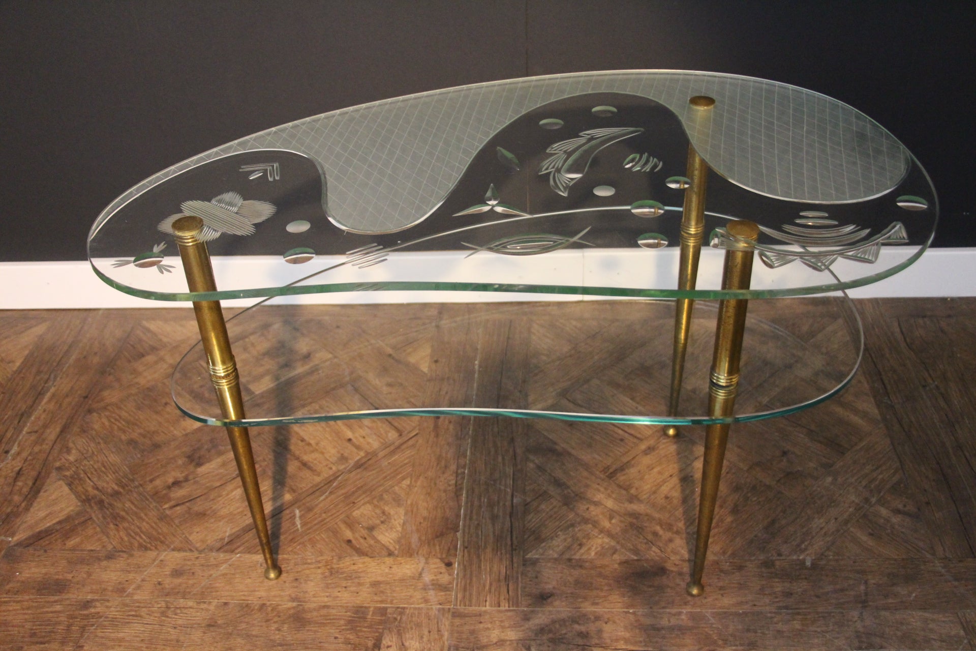 This very nice mid century glass top side table or coffee table features a free style shape and rests on 3 brass legs. It has got 2 glass tiers to provide extra storage.
Its glass top is a real work of art. Indeed, it features all hand engraved