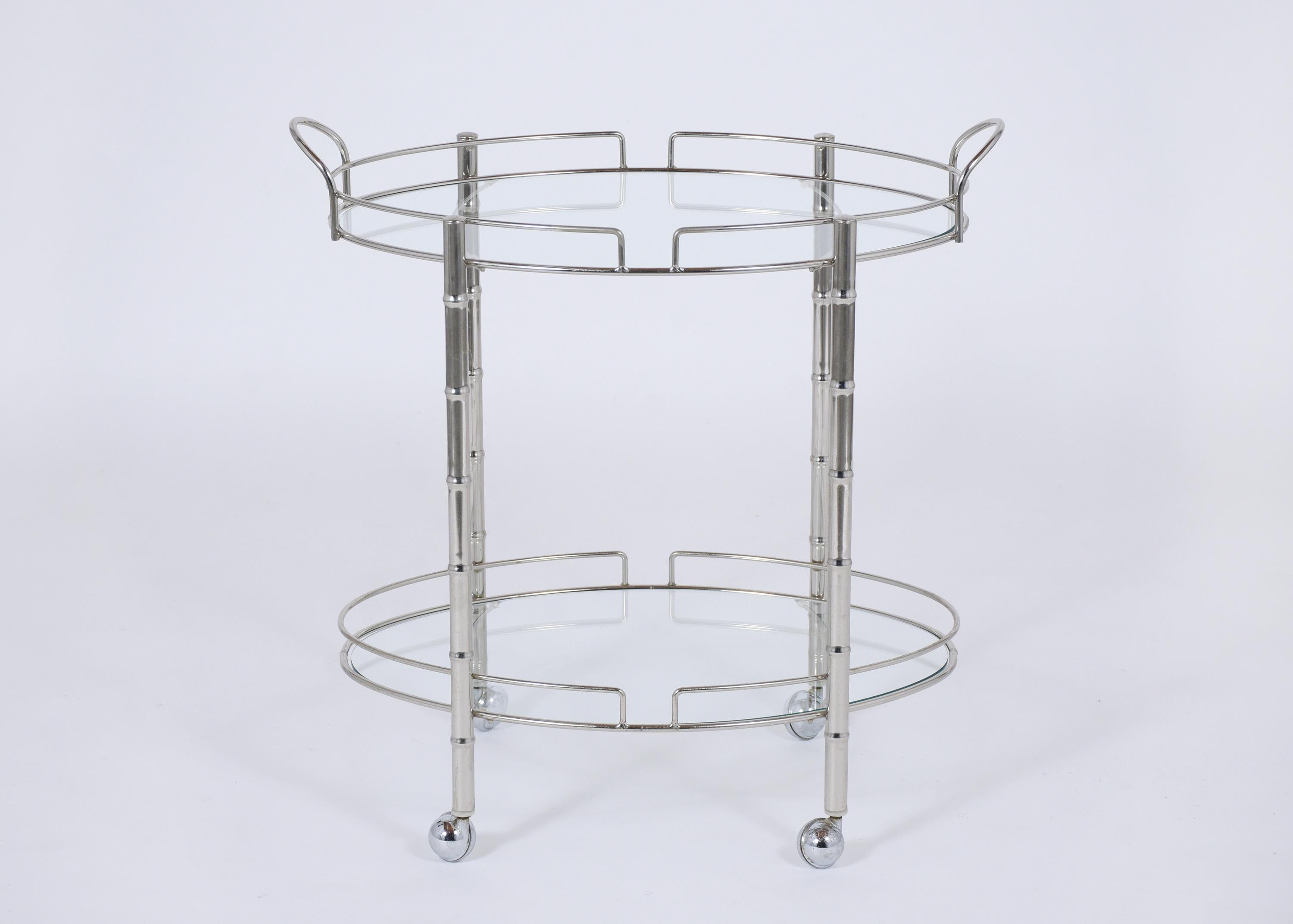 An extraordinary vintage mid-century bar cart is in good condition hand-crafted out of steel. This piece is eye-catching features a two-tier oval design clear glass tops with a flat polished finish in great condition supported by four faux bamboo