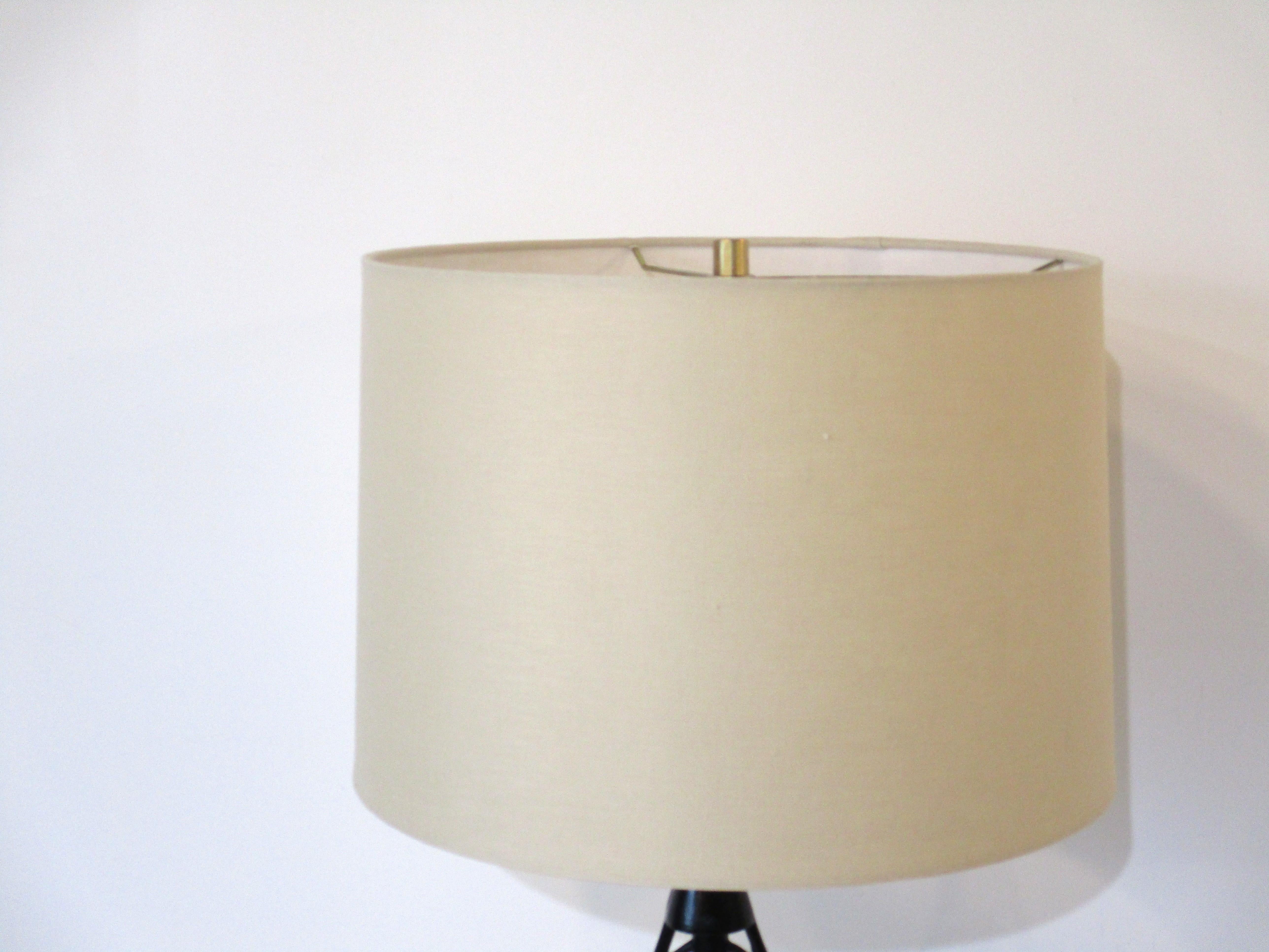 A welded black iron sculptural body table lamp having a fish bone motif sitting on a wood base with brass detail. Topped with a light beige linen drum shade and brushed brass final, designed in the manner of Frederick Weinberg and Heifetz.
