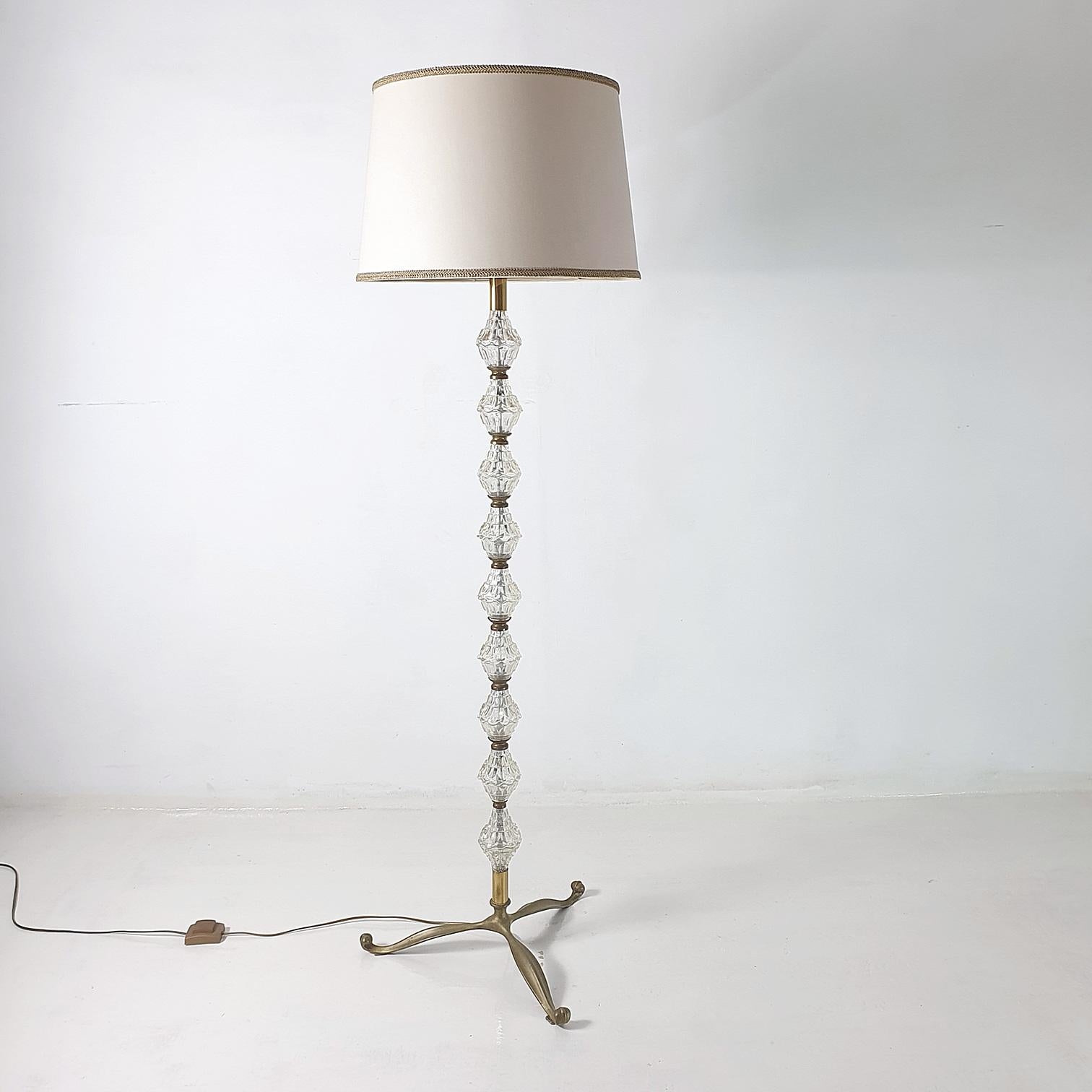 An elegant mid-century floor lamp produced in Italy in the 1950's in glass and metal crowned with an off white lamp shade with gold trimming. The shade is tan colored inside giving a warm glow when lit. Uses an E40 Edison bulb. Wired for European