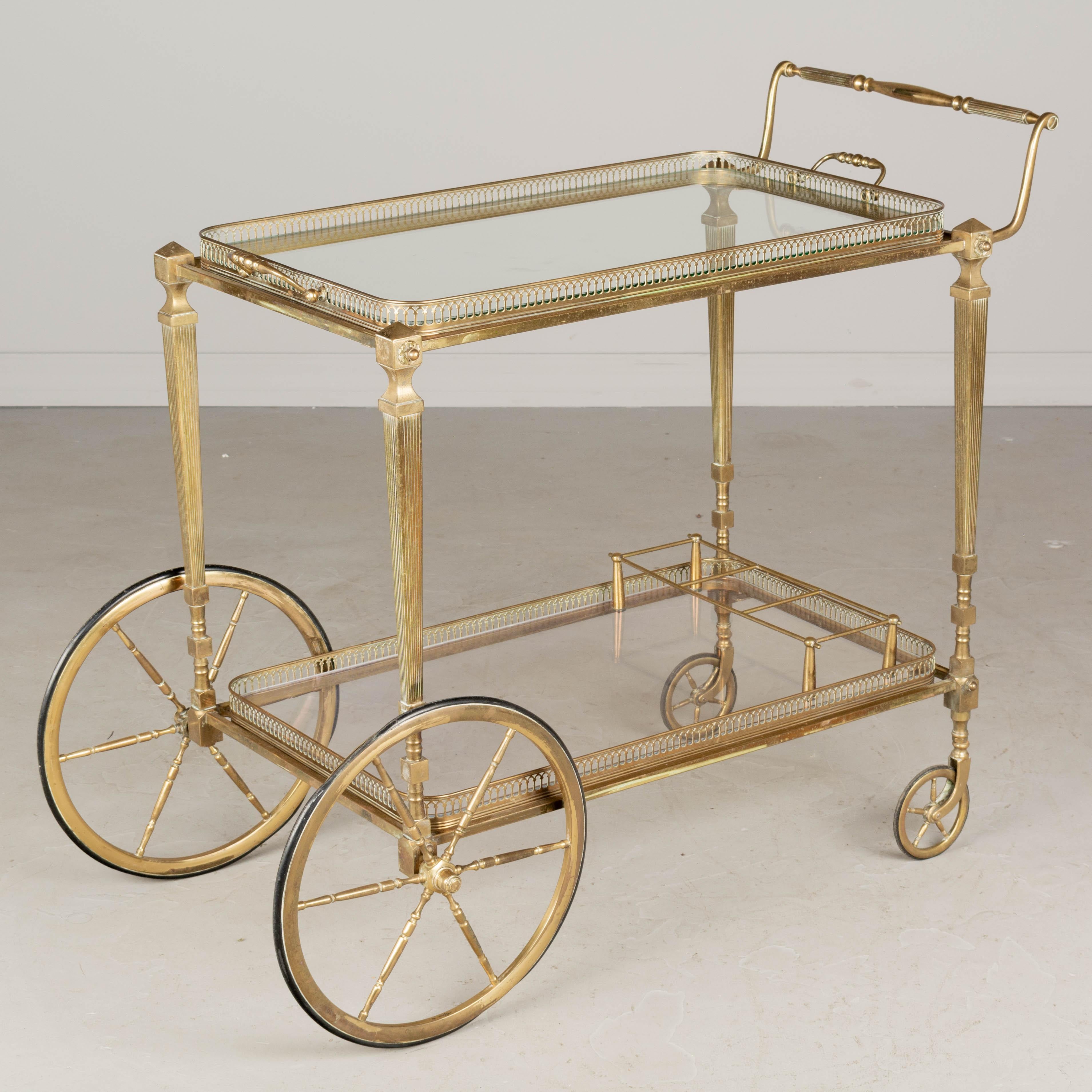 A Mid-Century Modern French brass bar cart in the style of Maison Jansen. The top shelf in glass and has handles and may be removed for use as a serving tray. The bottom shelf has a bottle rack and is plexiglass (as found). Nice patina. One of the