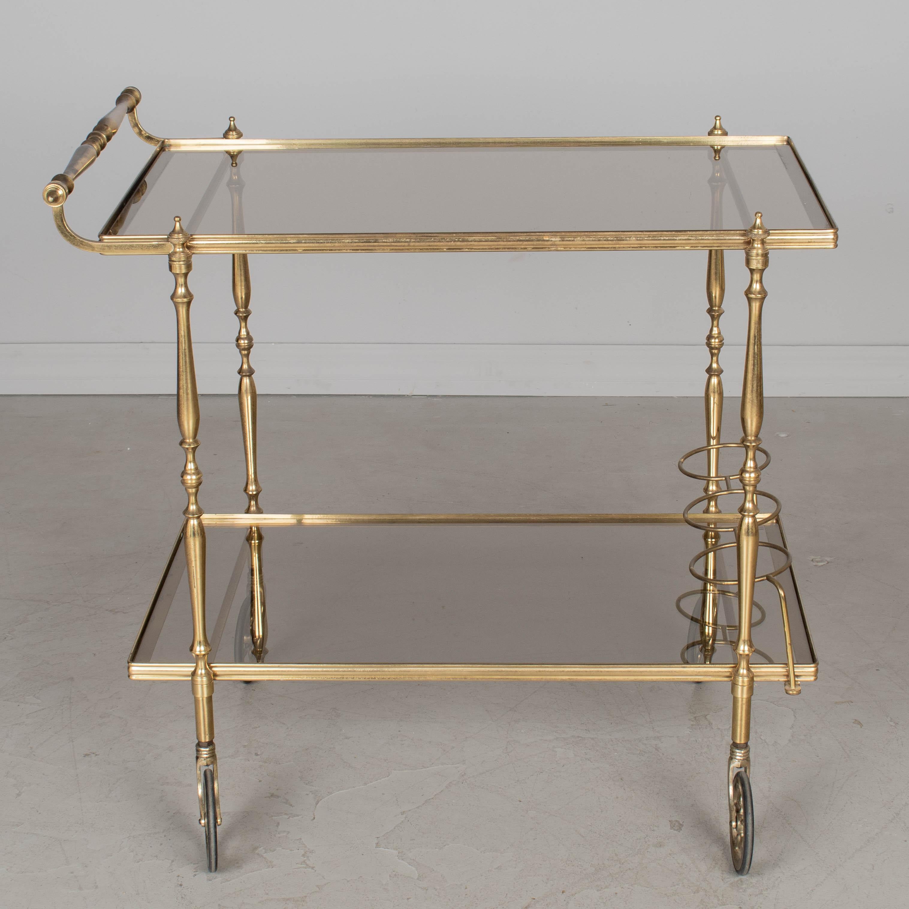 Chariot de bar en laiton et verre fumé, de style Maison Jansen, datant du milieu du siècle dernier. L'étagère inférieure est équipée d'un porte-bouteilles. Circa 1950s
29 