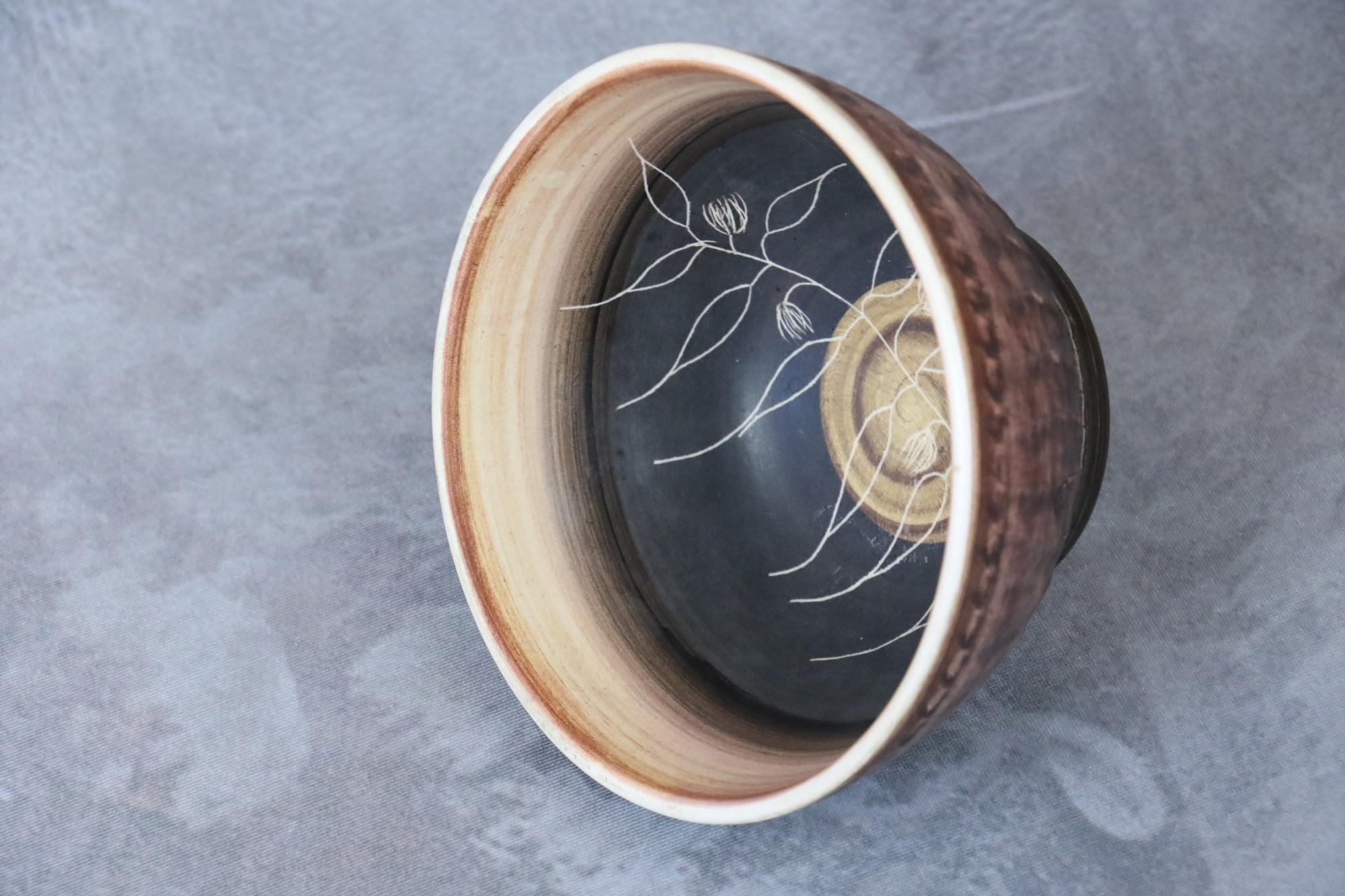 The bowl has earthy, black and white colours and is matt in appearance. 
Vegetal patterns are drawn on the inside of the bowl. 
The piece is signed 