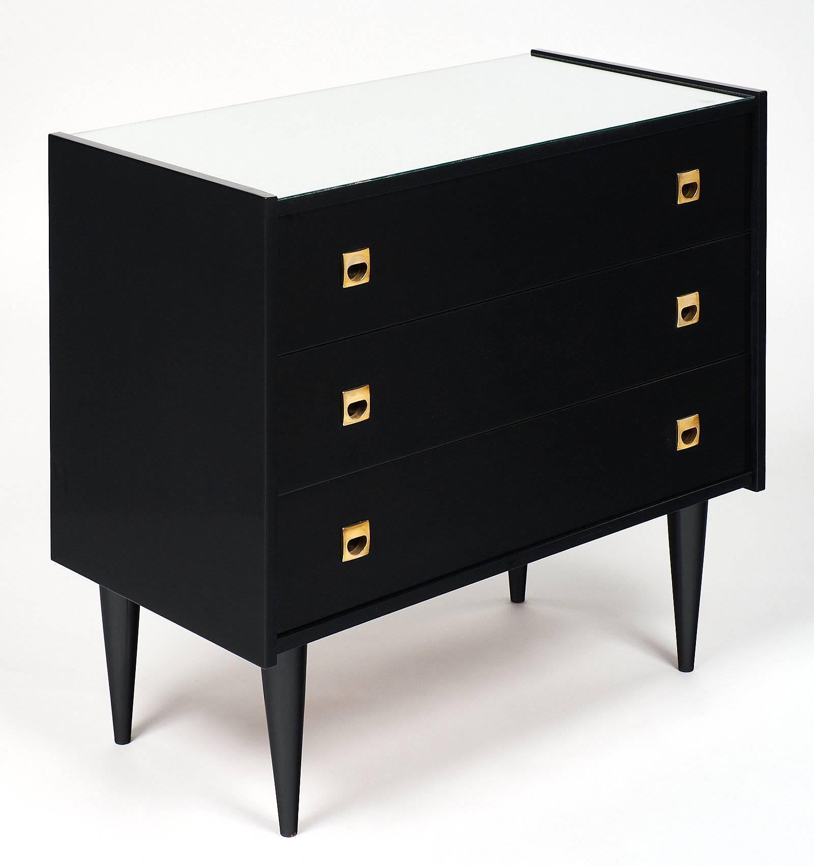 A French midcentury chest of drawers in ebonized cherrywood, finished with a high gloss French polish and a mirrored top. The three drawers each feature two inserted solid gilt brass handles. We love the quintessential Mid-Century Modern tapered