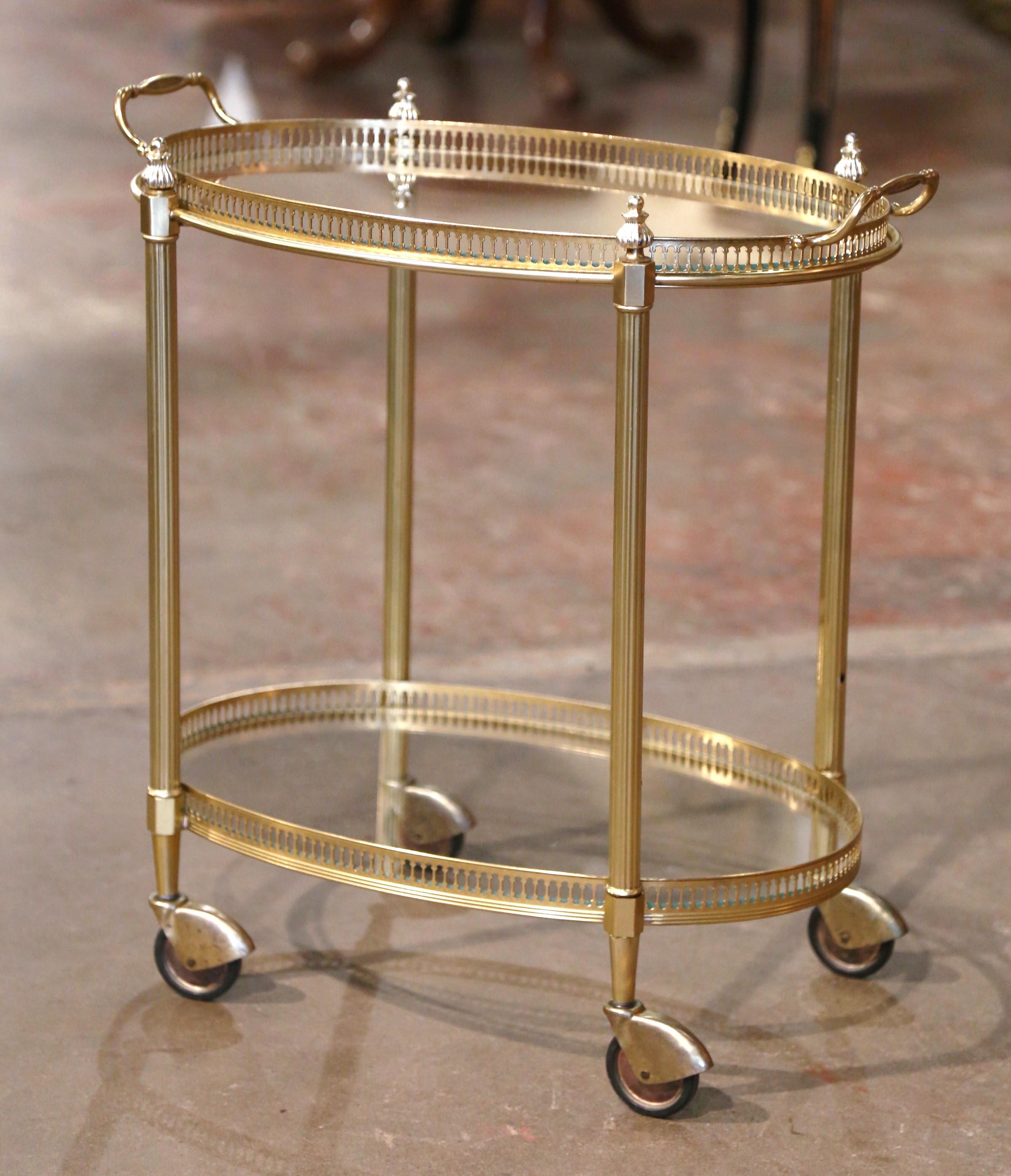 This elegant vintage rolling bar cart was created in France, circa 1960. Built in brass and oval in shape, the two-tier plateau trolley stands fluted legs topped with decorative finials, and ending with small casters at the base. The top deck,