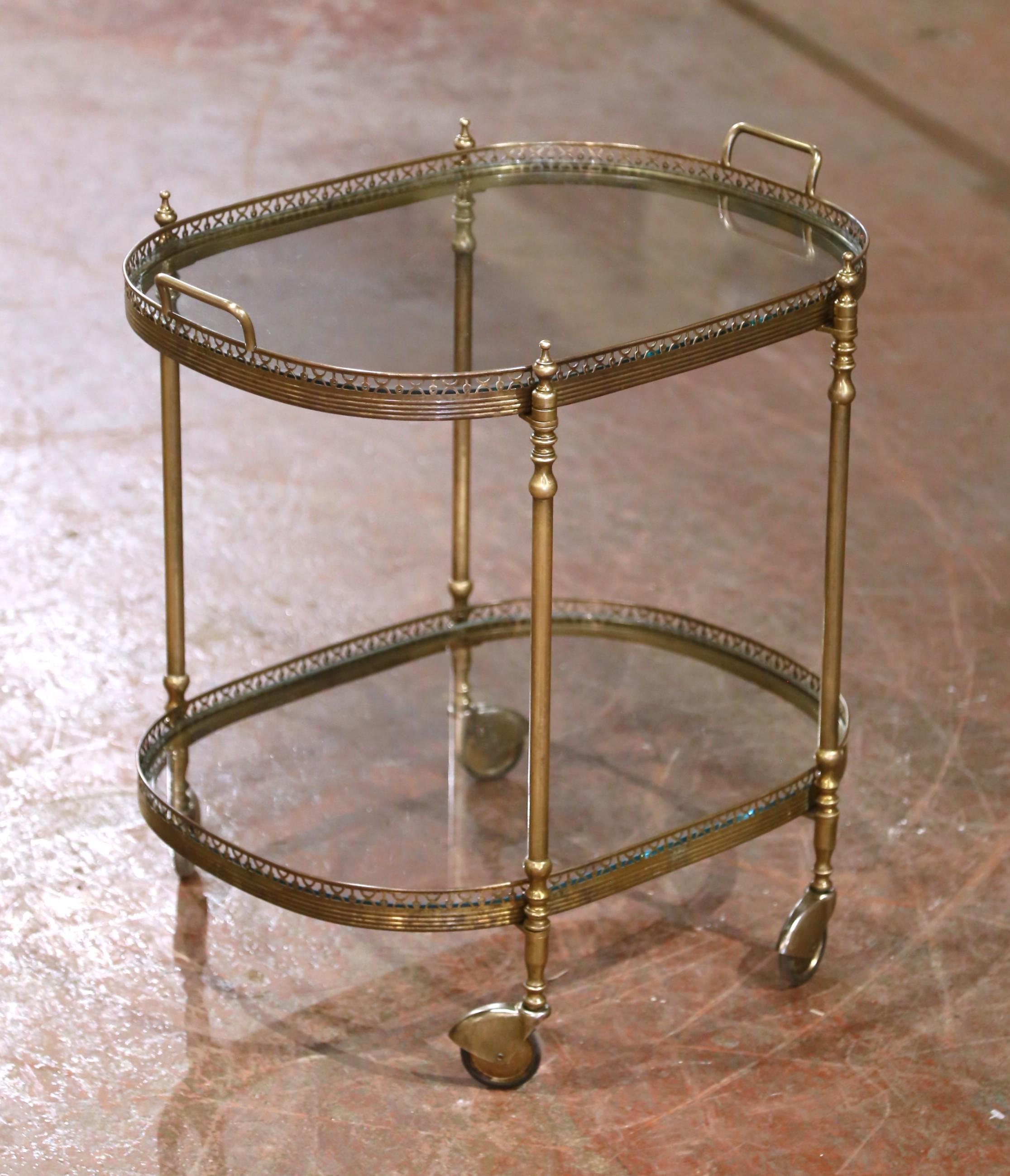 This elegant two-tier bar cart was crafted in France circa 1960 and embodies the sleek sophistication of mid-century design. Oval in shape and built from gilt brass, the vintage service trolley features fluted legs topped with decorative finials and