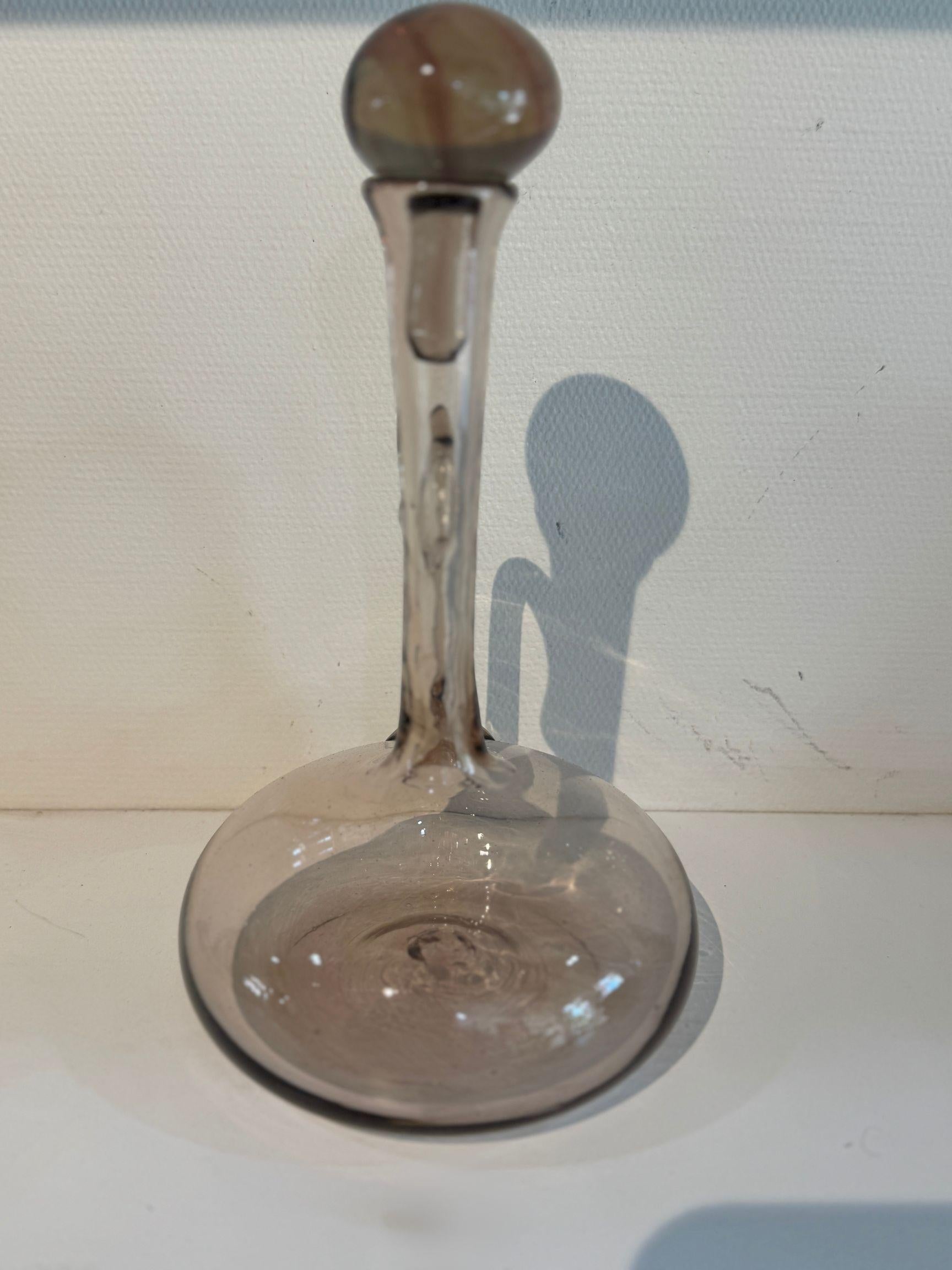 Un decantador de vino francés de mediados de siglo de bellas proporciones, soplado a mano en vidrio marrón grisáceo ahumado y rematado con un llamativo tapón esférico. Con su cuello alargado, su asa curvada aplicada y su amplia base redondeada, esta