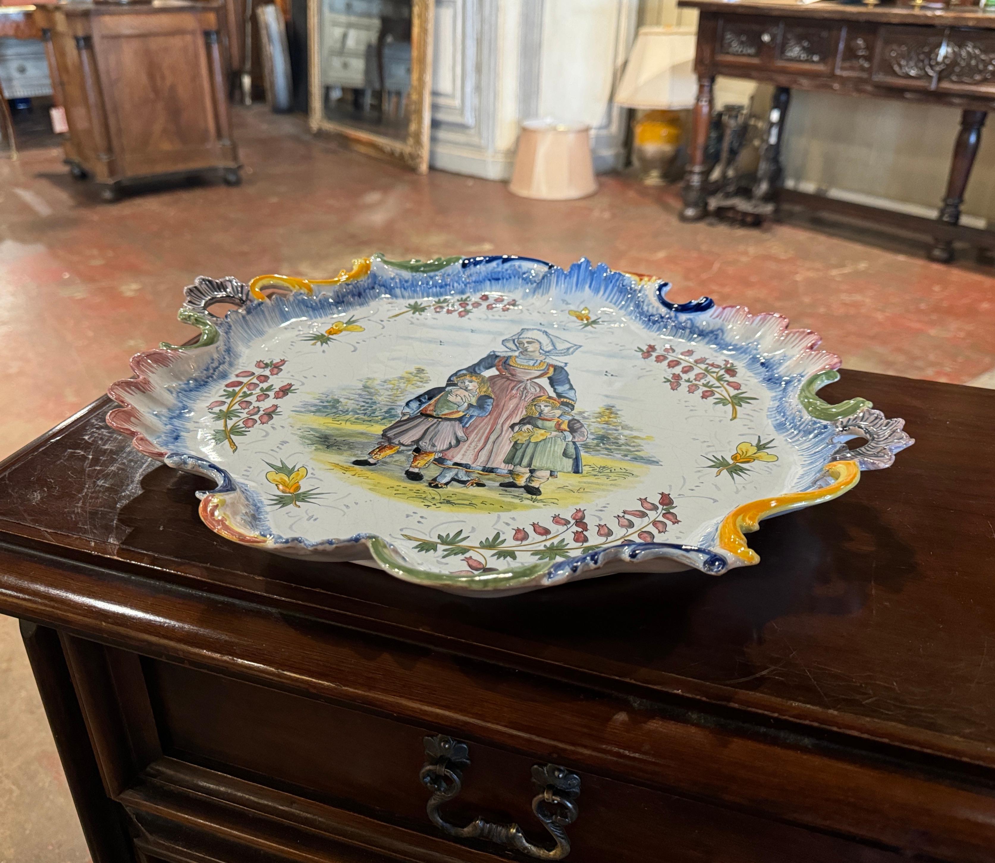 Décorez un mur ou une étagère avec ce plat antique coloré. Créée en Bretagne (France) vers 1950, cette assiette en céramique peinte à la main est de forme ronde et habillée de petites poignées sur les bords festonnés. Le grand plateau présente une