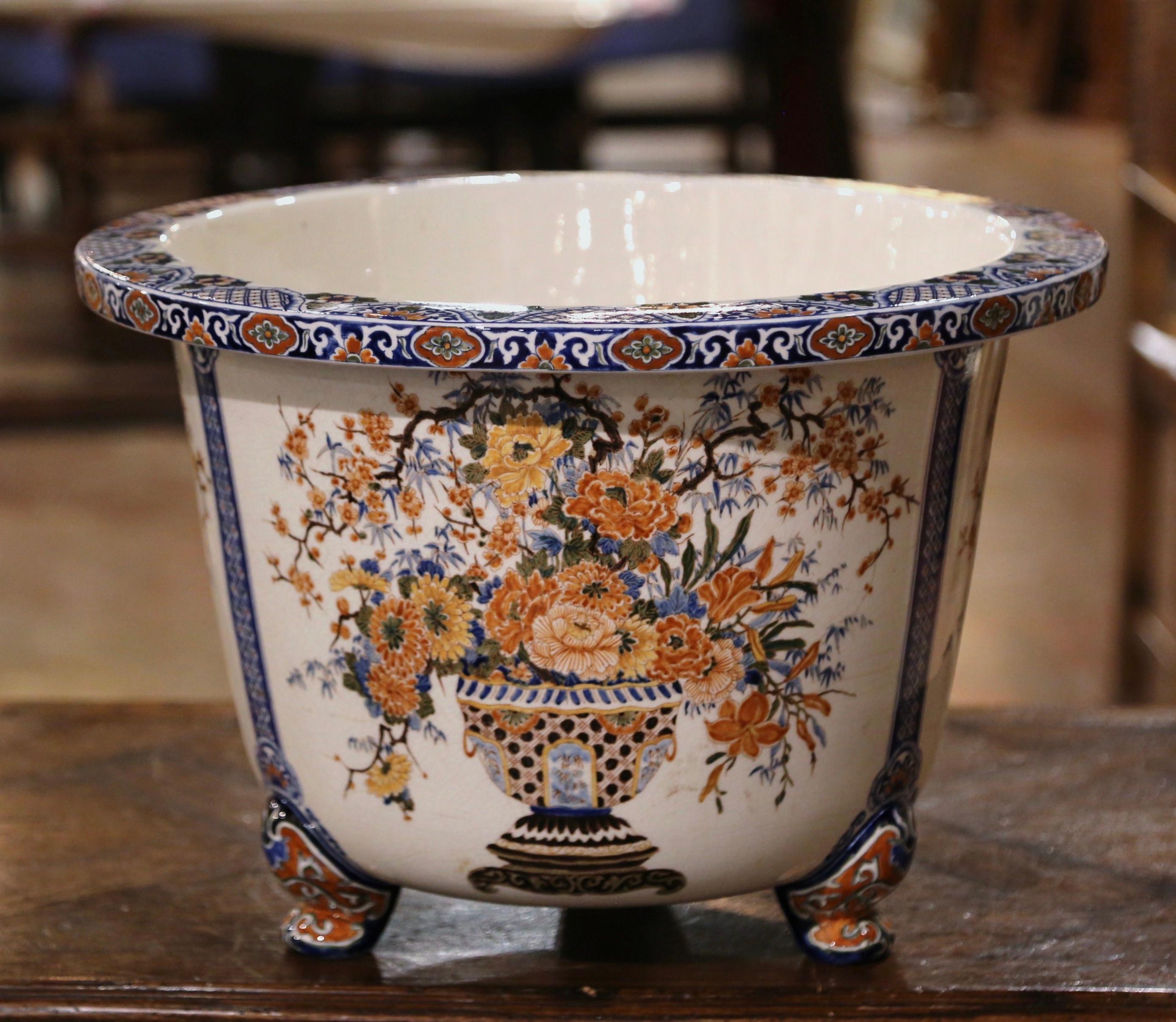 This important and colorful antique cache pot was crafted in Normandy, France, circa 1950. Standing on three dolphin form feet, the large porcelain planter is decorated with three floral medallions around the perimeter, and embellished with a blue
