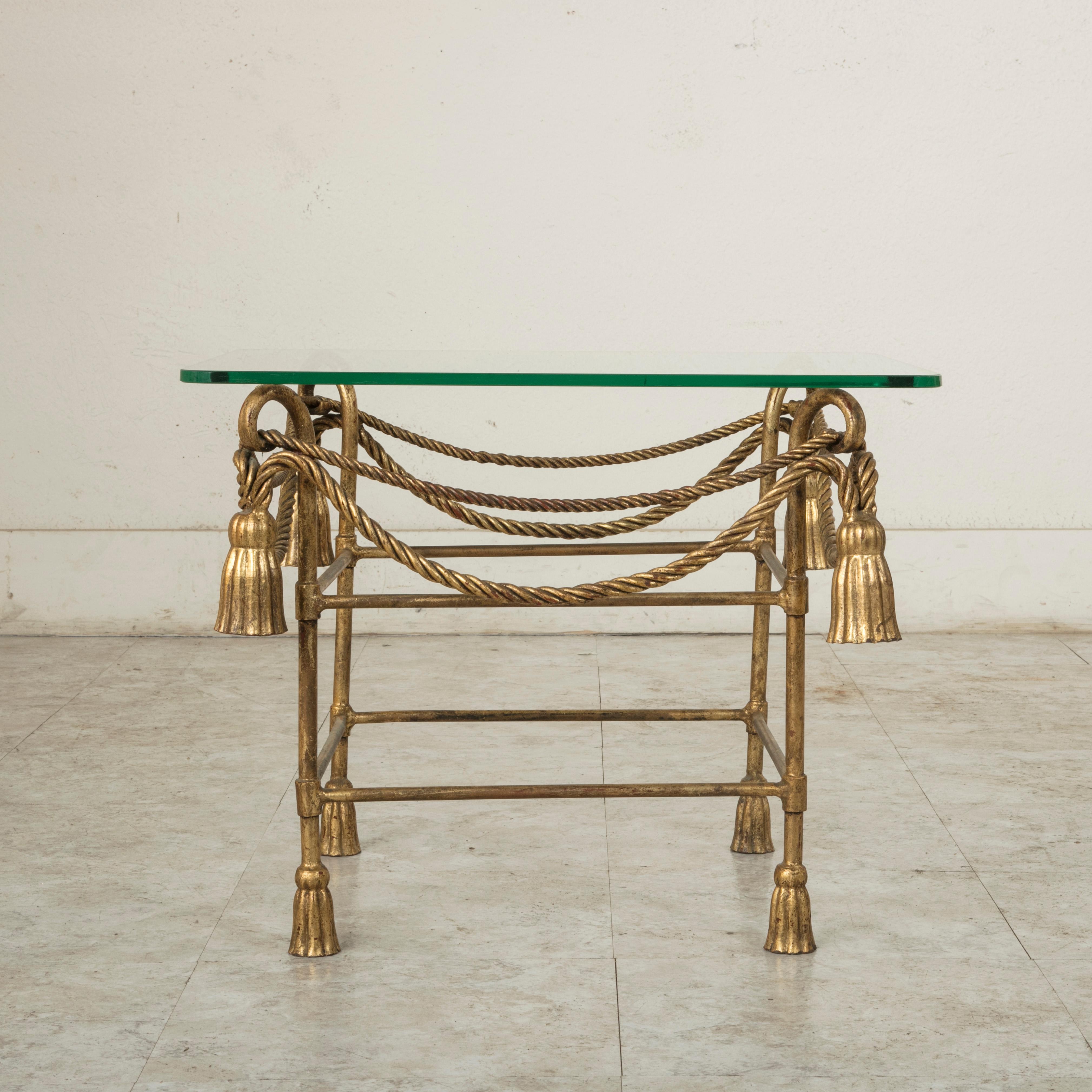 Attributed to French designer Jean-Charles Moreux (1889-1956), this midcentury gilt iron side table or end table features rope swags and tassels that hang from its scrolling top. The piece is capped with a glass top and rests on additional tassel