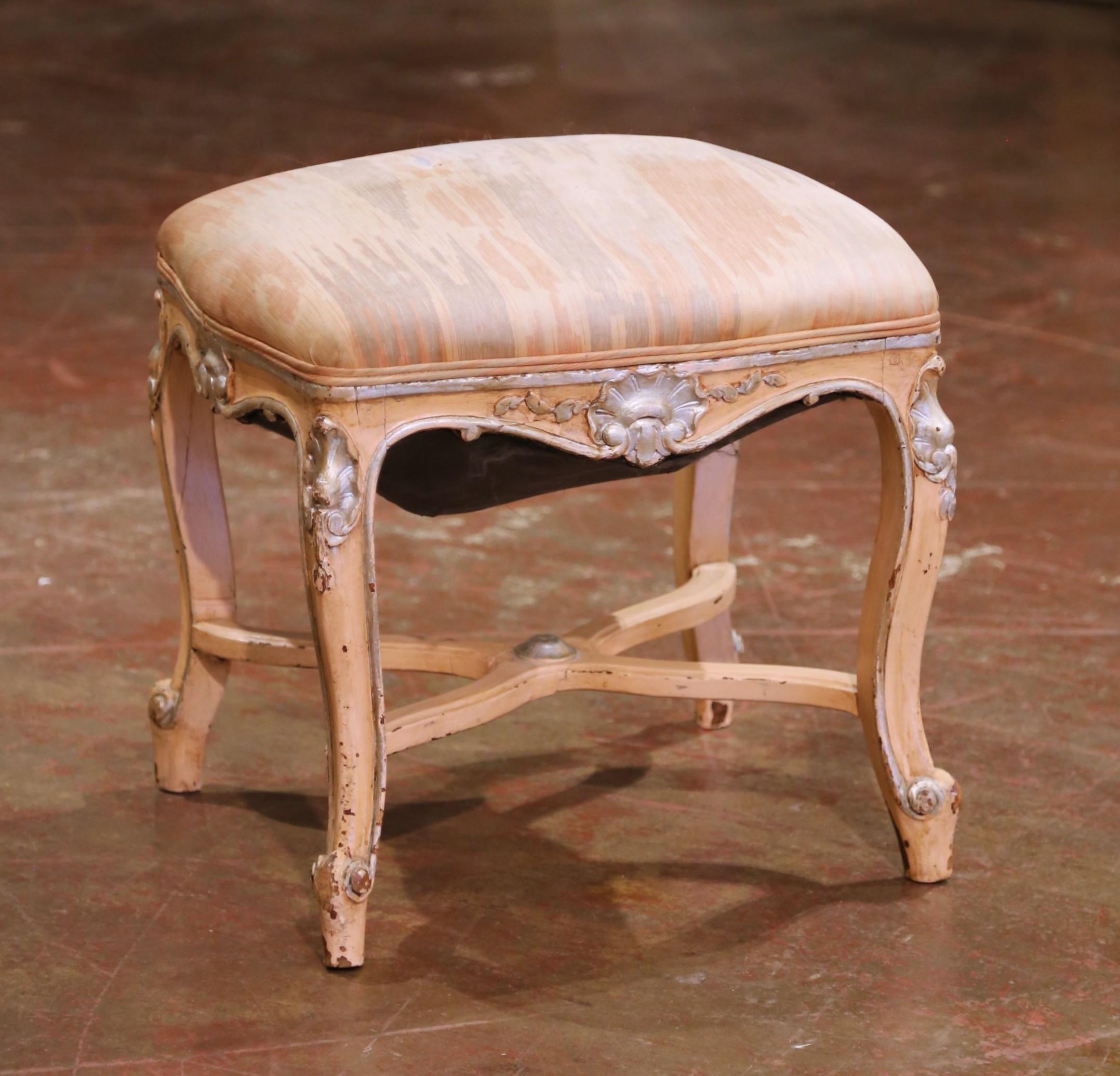 Dieser dekorative französische Hocker aus der Mitte des Jahrhunderts, der um 1960 im Louis-XV-Stil gefertigt wurde, zeichnet sich durch einen handgeschnitzten Buchenholzrahmen mit einer sanften cremefarbenen Lackierung aus, die durch Muschel- und
