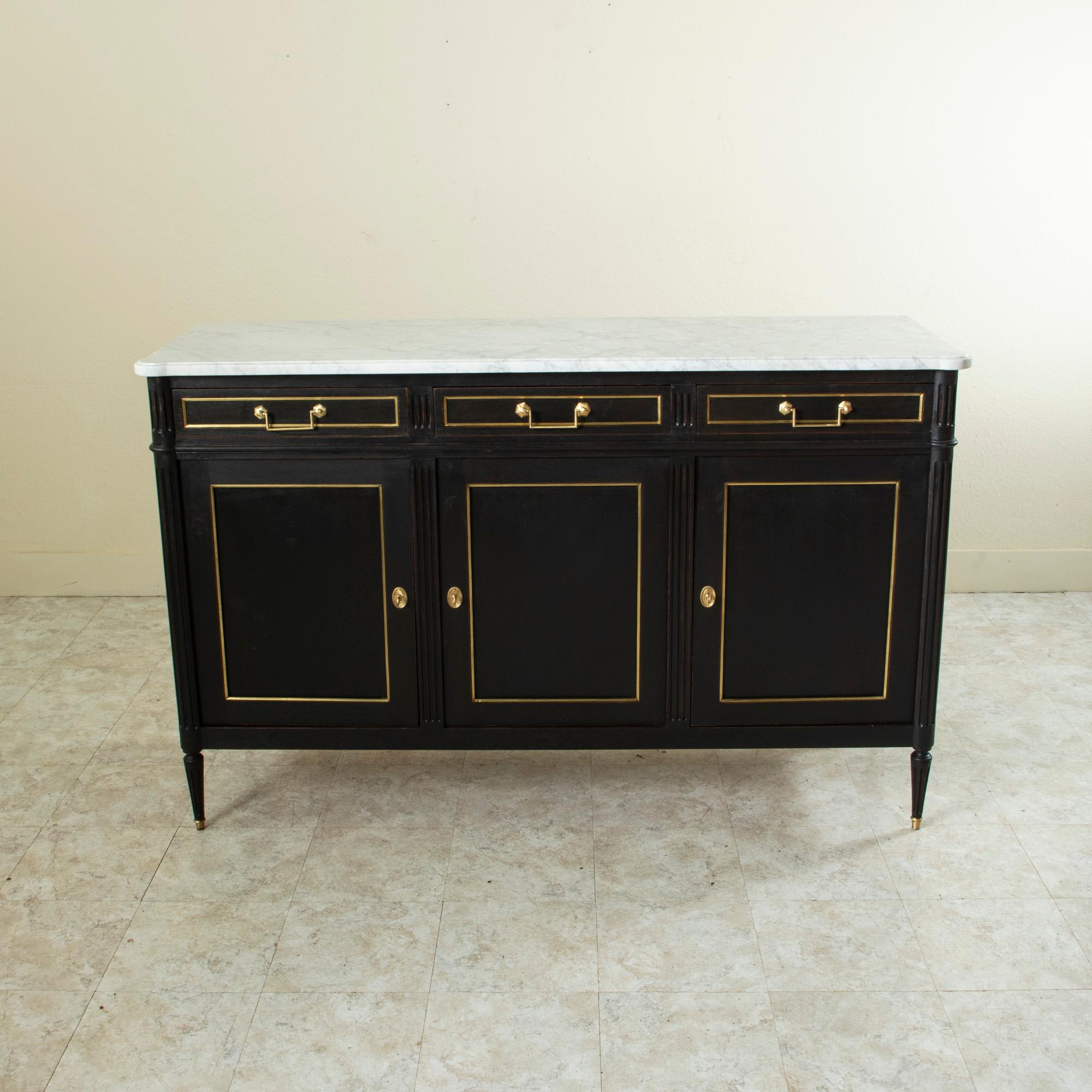This French Louis XVI style enfilade or sideboard from the mid-20th century features a black painted mahogany cabinet with a white marble top. Stunning bronze banding outlines each door and drawer, while fluted columns define the facade. Its