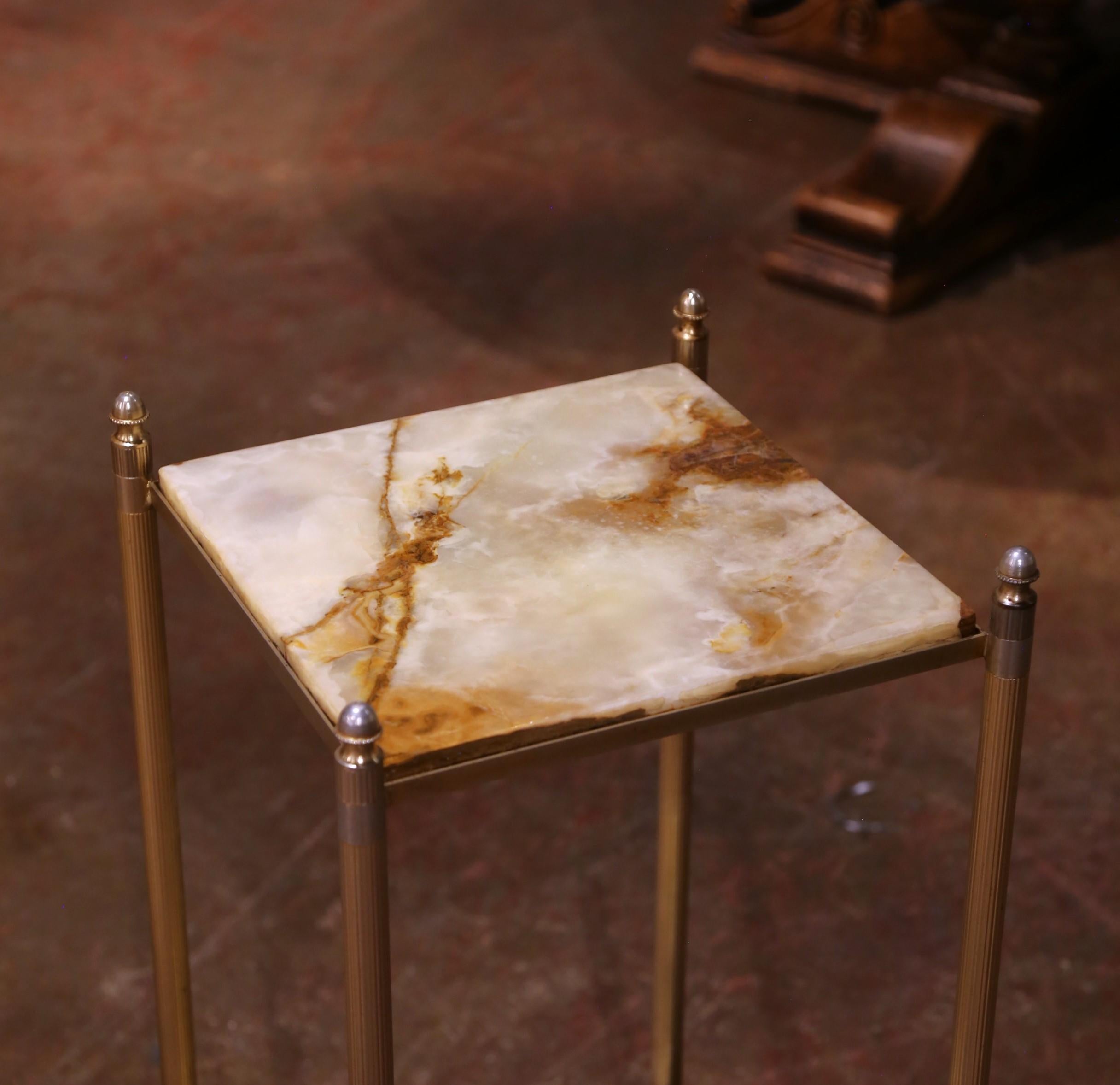 The gilt-brass frame features finely fluted legs, decorative beaded collars, and tapered feet, each capped with neoclassical finials. Both tiers have square tops inset with light onyx panels, their creamy beige ground enlivened by warm honey,