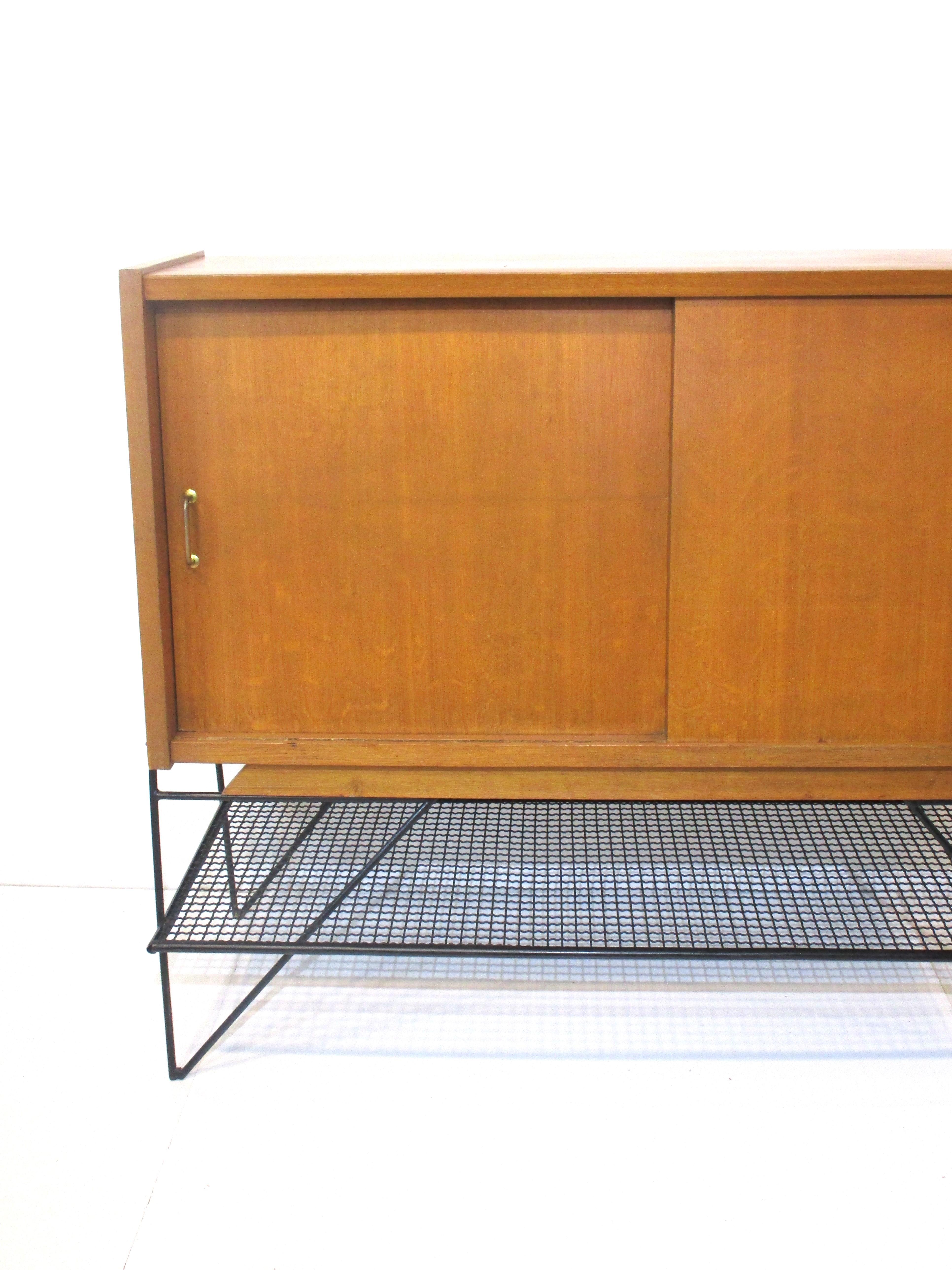 A French oak sliding two door credenza / cabinet with brass pulls one fixed shelve , storage and a small drawer . The cabinet tappers at an angle upwards and sits on a sculptural black iron base with grated lower shelve platform . A nice piece
