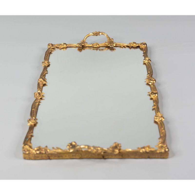 A stunning French gilt brass rectangular mirror vanity or bar tray, circa 1940. This beautiful ormolu tray is decorated in a French Rococo style with roses, scrolling leaves, and ornate handles in a lovely gilt patina. It would be gorgeous for
