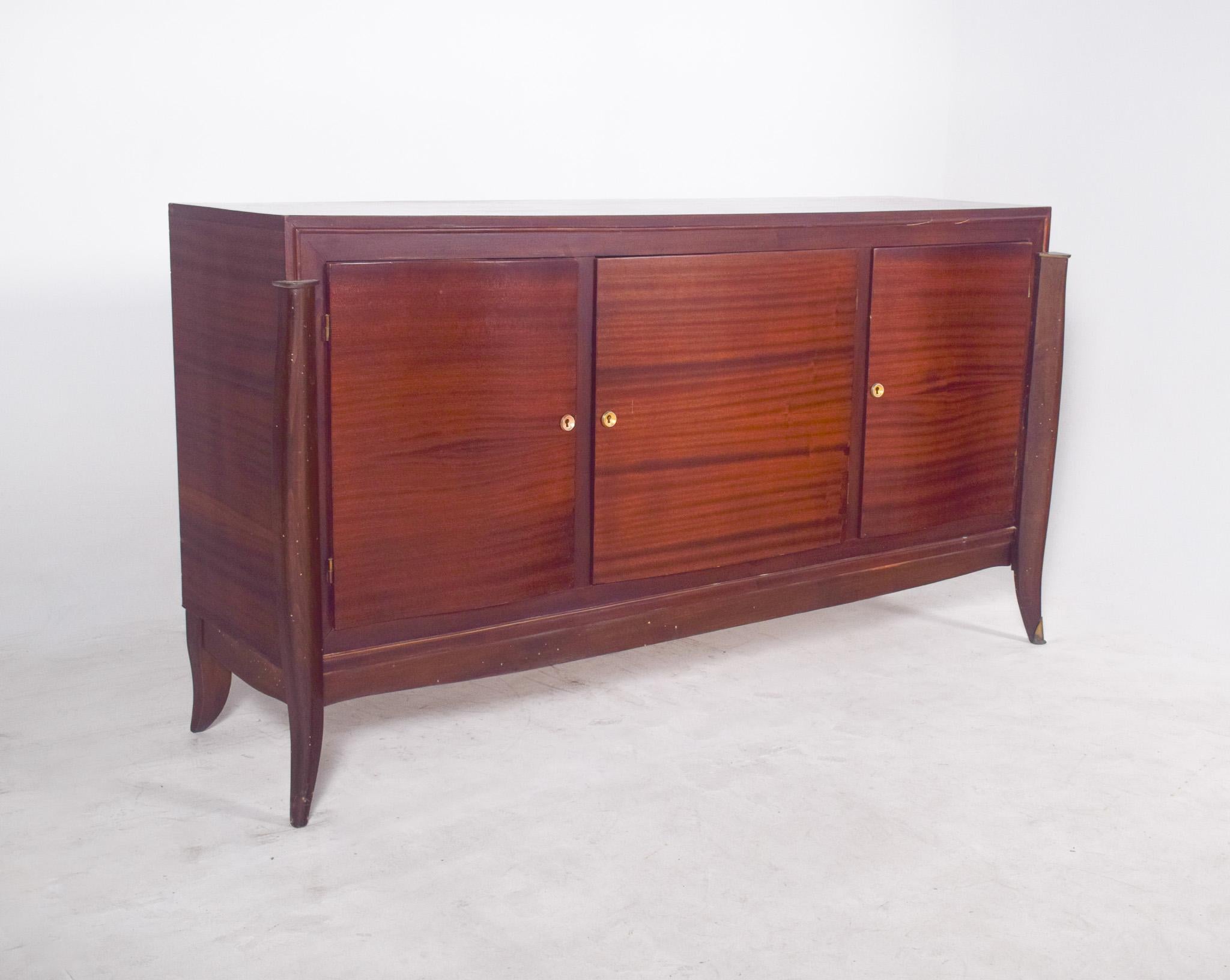 Buffet à trois portes des années 1950. Les pieds, hauts et arqués, se prolongent à l'avant jusqu'au sommet de la structure, créant ainsi un cadre vertical qui entoure le meuble et se termine par de délicates pointes métalliques. Ce détail vertical