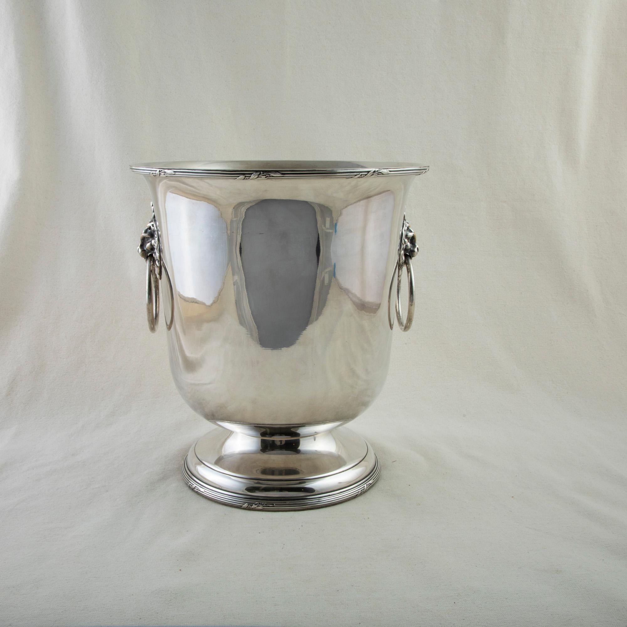 This mid twentieth century French silver plate champagne bucket features a lion's head on each side with a drop ring handle. The fluted upper rim is detailed with leaves, and the bucket stands on a footed base. It is stamped with a hallmark on the