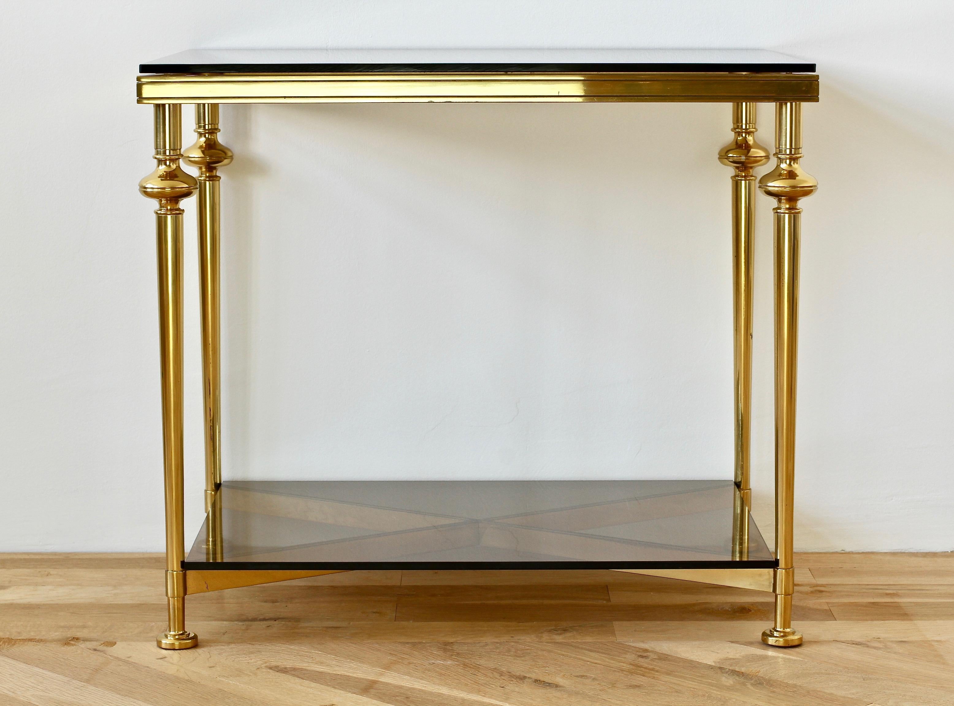 A wonderful side or end table in the style of Maison Jansen, circa 1970. Made from solid cast brass in the modern neoclassical style and featuring two dark toned smoked glass tabletops and crossed X-base. This delightful table, not dissimilar to