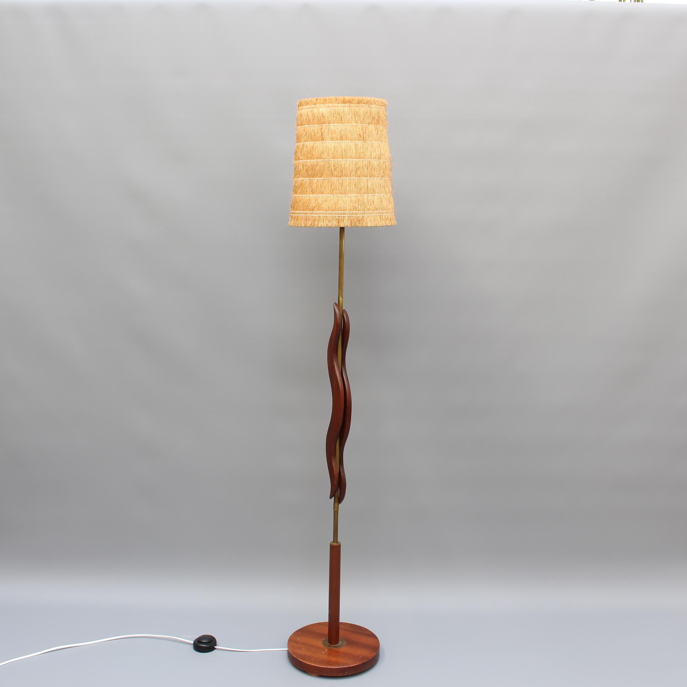 Midcentury French wooden and brass floor lamp (circa 1950s). A beautiful reddish wood intermittently covers the brass lamp pole which ascends to the stylish, raffia lampshade. The curved wood is reminiscent of the flame from a candle. The round