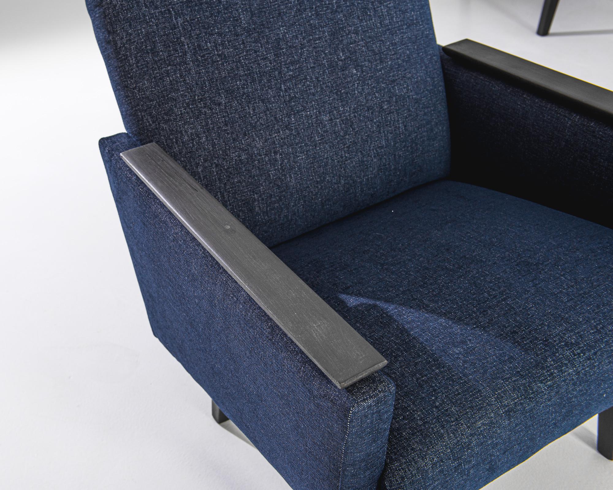 A pair of upholstered wooden armchairs from France, produced circa 1960. A smart set in a deep, dark, denim blue, featuring black patinated wooden armrests and legs. Chic slants create a sloping seat back and trapezoidal side panels in this
