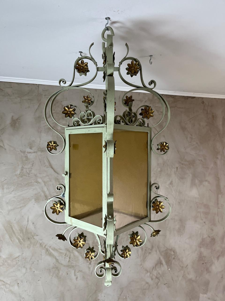Add a touch of timeless elegance to your interior or terrace with this beautiful hanging lantern in classic-style wrought iron. Painted in a soft patinated pale green, it features finely crafted golden floral details, offering a romantic and refined