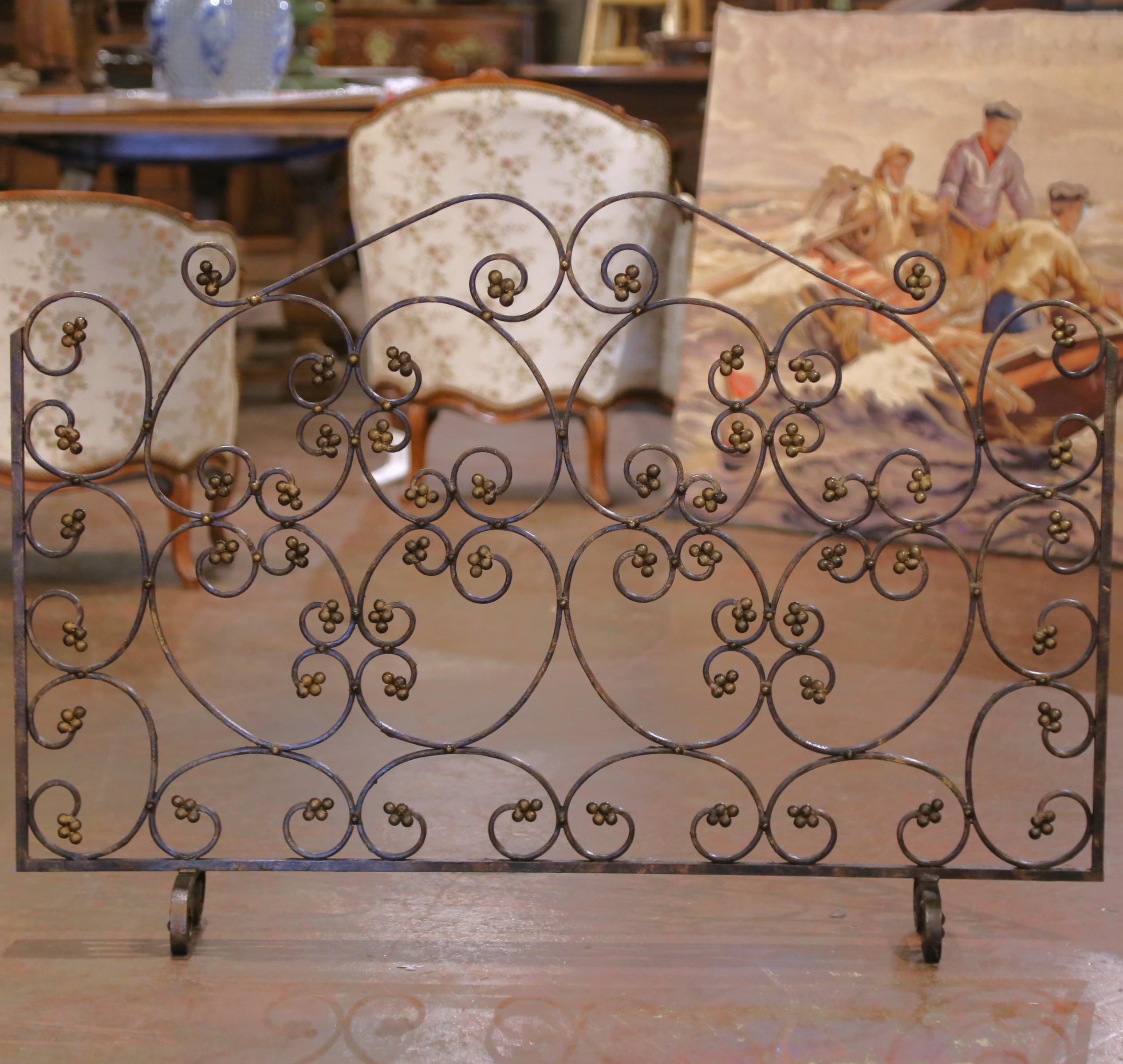 Crafted in France circa 1960, this elegant mid-century firescreen showcases an ornate scrolling design in hand-forged wrought iron. The openwork composition features symmetrical curves accented with clusters of gilt floral rosettes, lending a