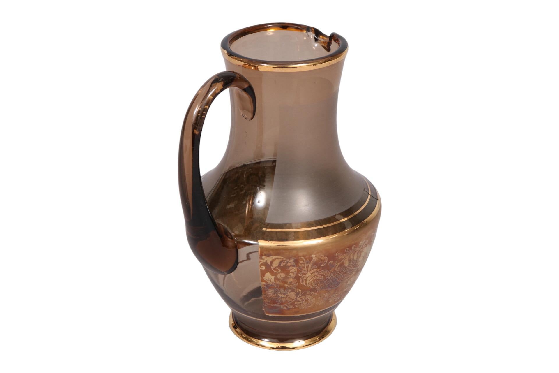 A midcentury frosted glass pitcher. The brown frosted glass has a thick gold band adorned with floral details around the middle and is edged with gold lines.