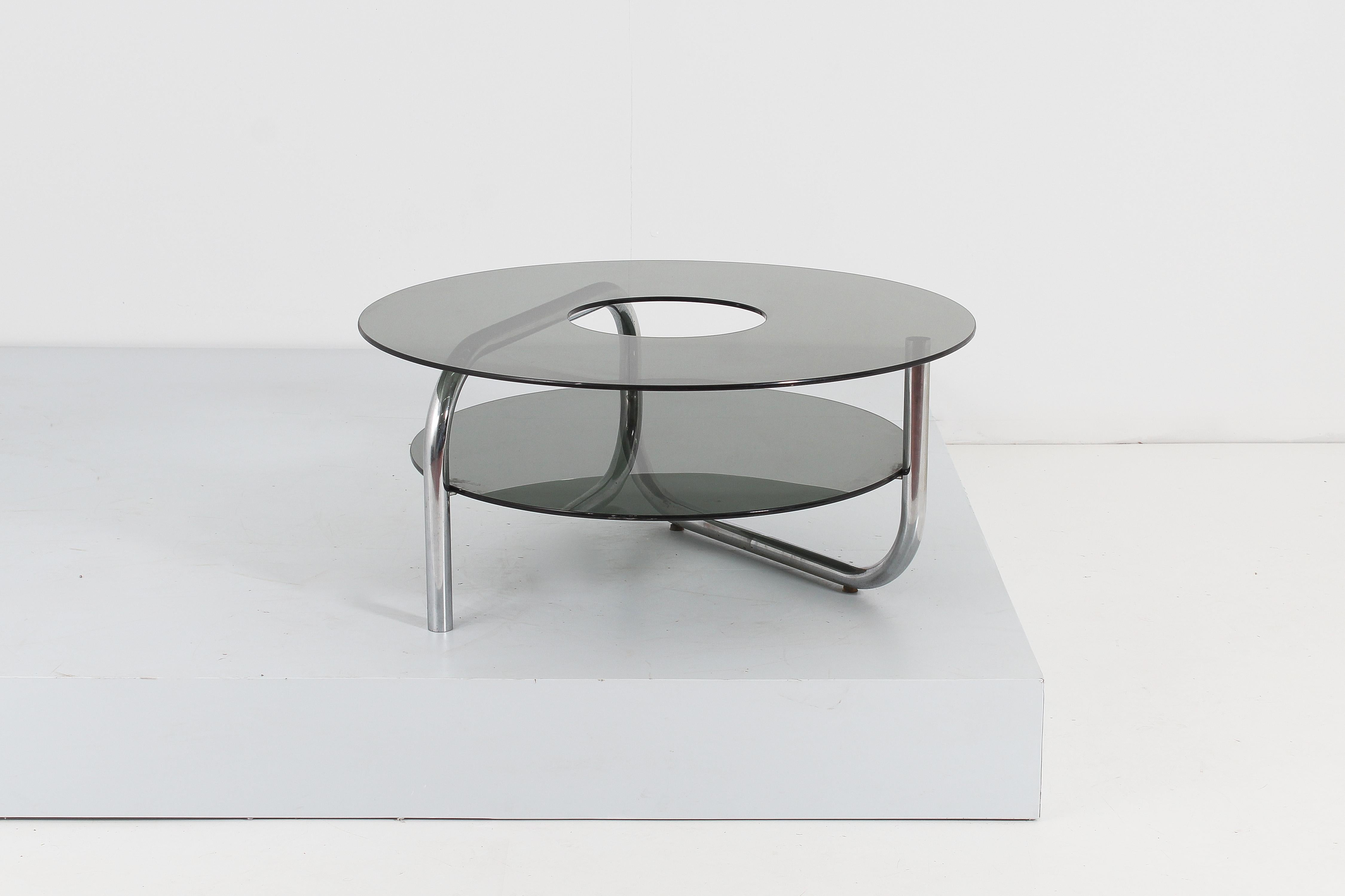 Stylish circular coffee table with double top in smoked glass with curved tubular support in chromed steel. The upper shelf, with a larger diameter than the lower one, has a circular hole in the center, to allow you to place bottles of liquor or