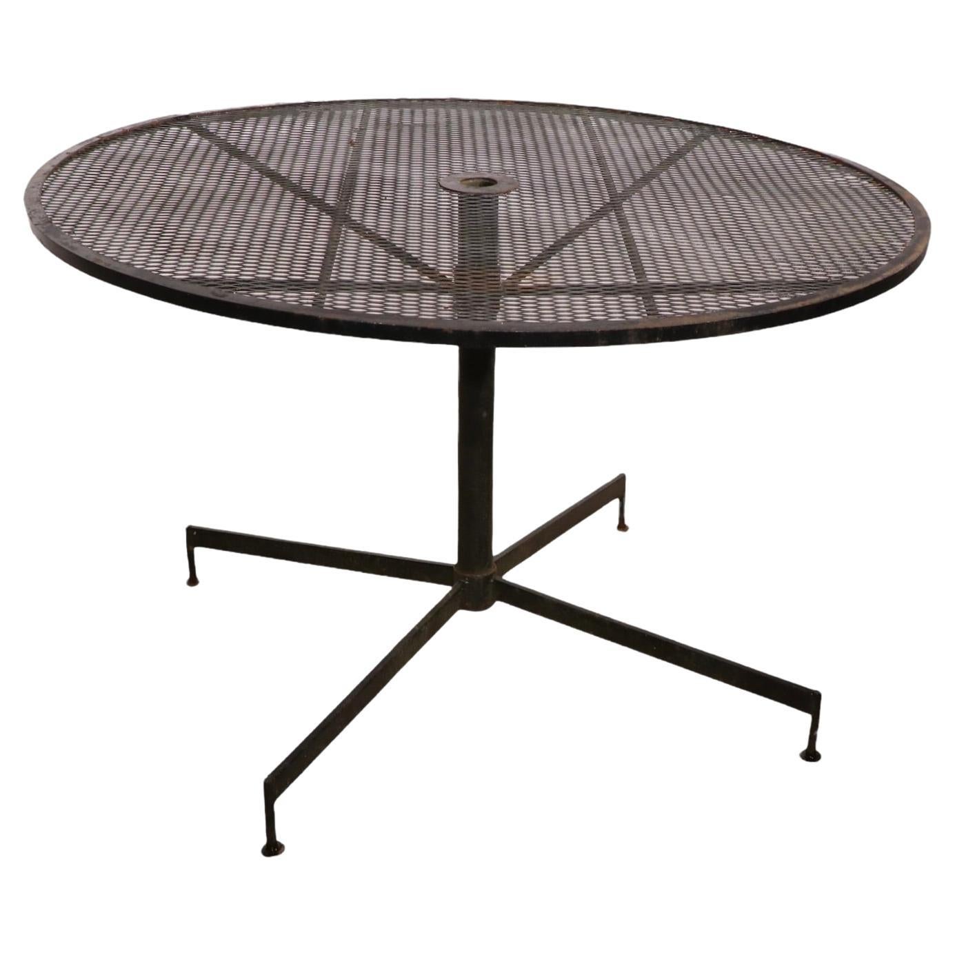 Chic architectural wrought iron and metal mesh garden, patio, poolside cafe dining table. The table features a circular metal mesh top, supported by an iron center pole, on a four leg star base. Construction reminiscent of Woodard or Salterini,