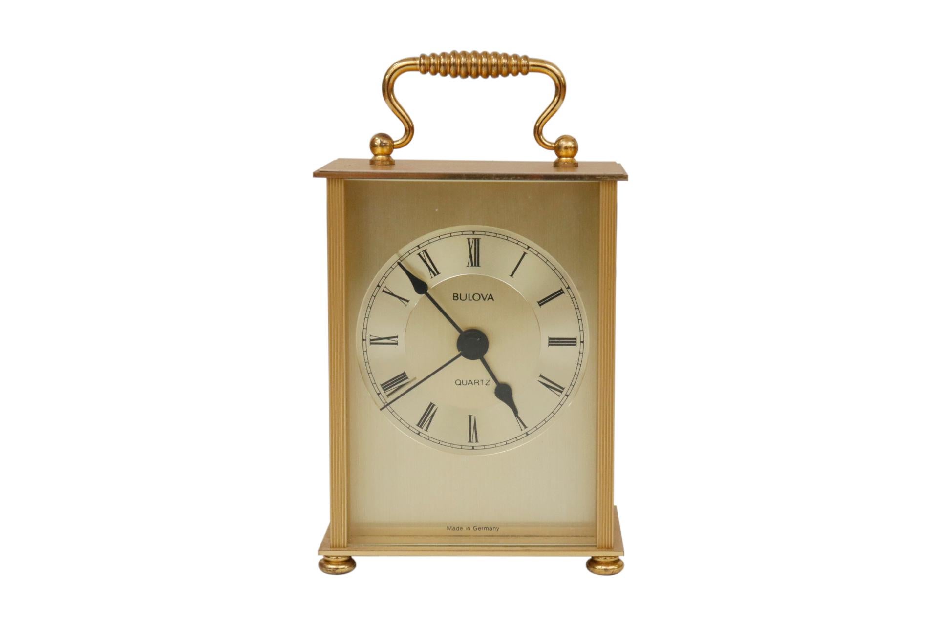 A traditional carriage clock made by Bulova. A simple clock face with Roman Numerals behind glass is set in a simple gold box topped with a bail handle. Engraved with three stripes down each side and finished with small bun feet.