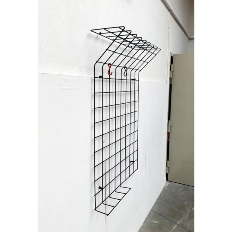 Simple wardrobe made of coated metal grille. The upper and part of the lower area are shaped into a shelf. The wardrobe is installed with integrated angles at a distance on the wall. The three colored original hooks can be moved as