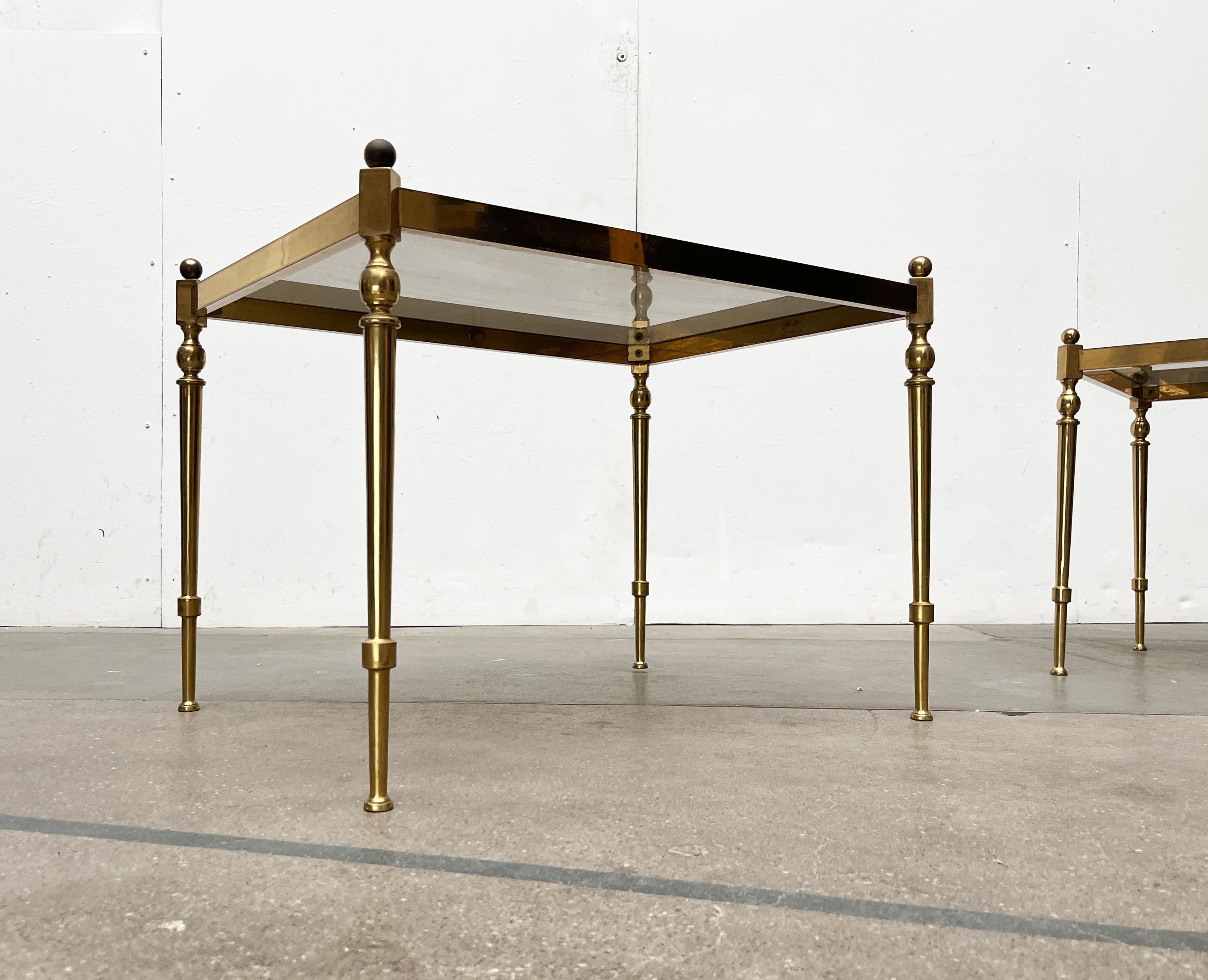 Deux tables d'appoint de style néoclassique datant du milieu du siècle dernier.

Les tables se composent d'un cadre en laiton et d'une surface en verre fumé. La structure solide forme un large cadre rectangulaire en haut qui entoure le plateau de la