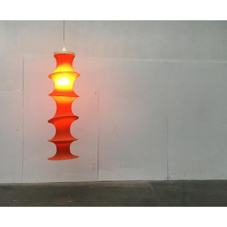 Lampada a sospensione Mid Century Tube di colore arancione del produttore di Luminaire Aro.
 
 un tessuto elastico arancione teso su un anello di plastica che funge da diffusore. Gli anelli sono flessibili e possono essere spostati manualmente, in